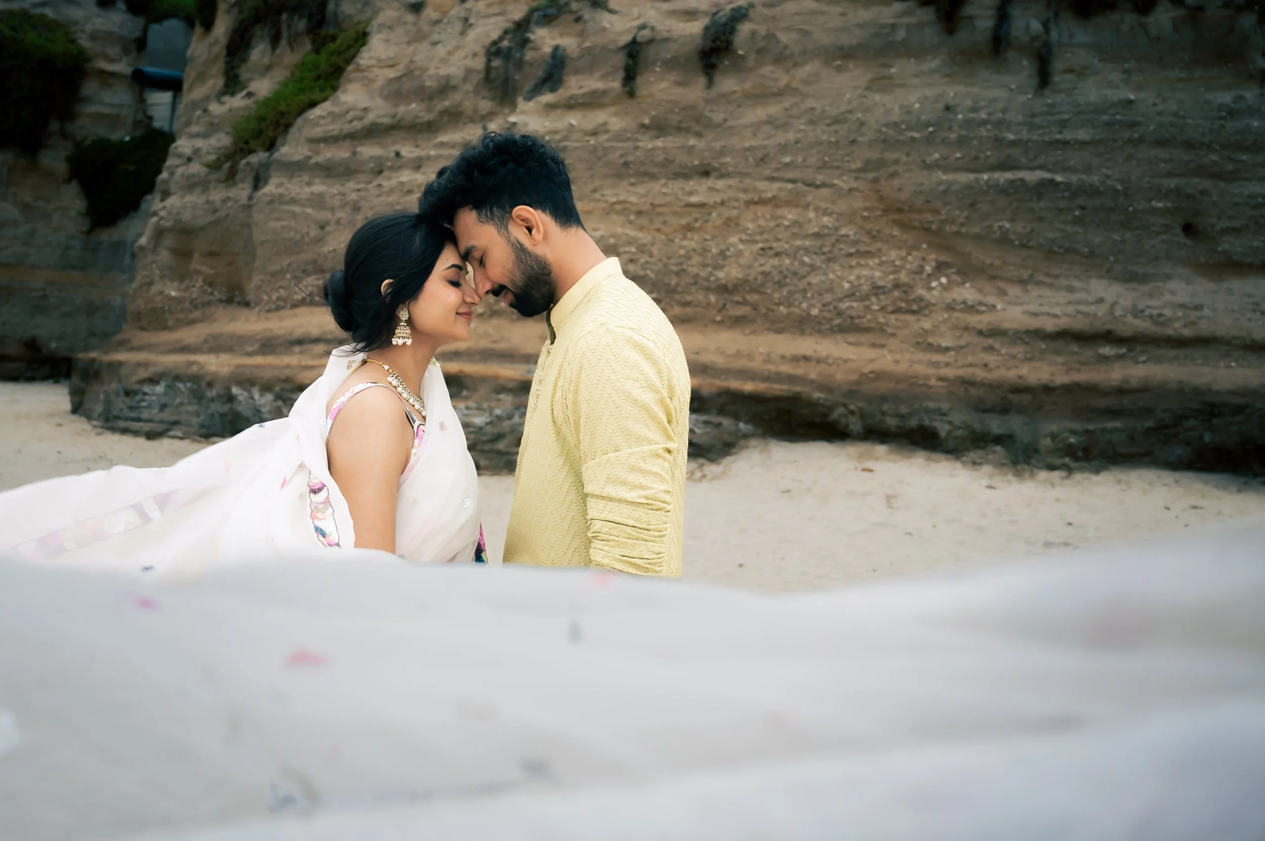 A couple dressed in traditional Indian attire sharing a romantic moment on the beach, with their foreheads touching and eyes closed.