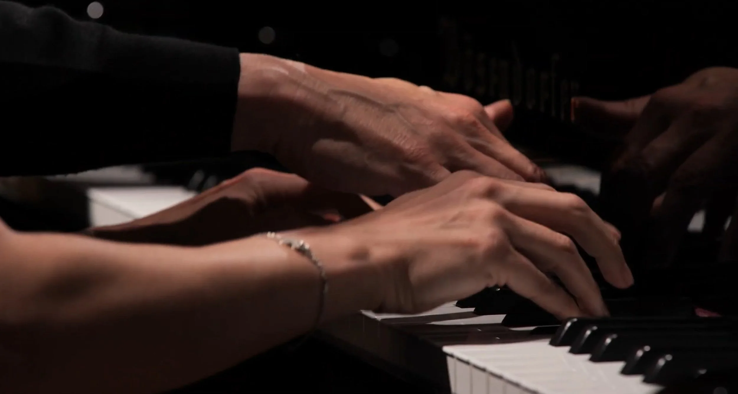Mains de deux personnes jouant du piano, une main plus âgée et une main plus jeune, avec un bracelet en argent.