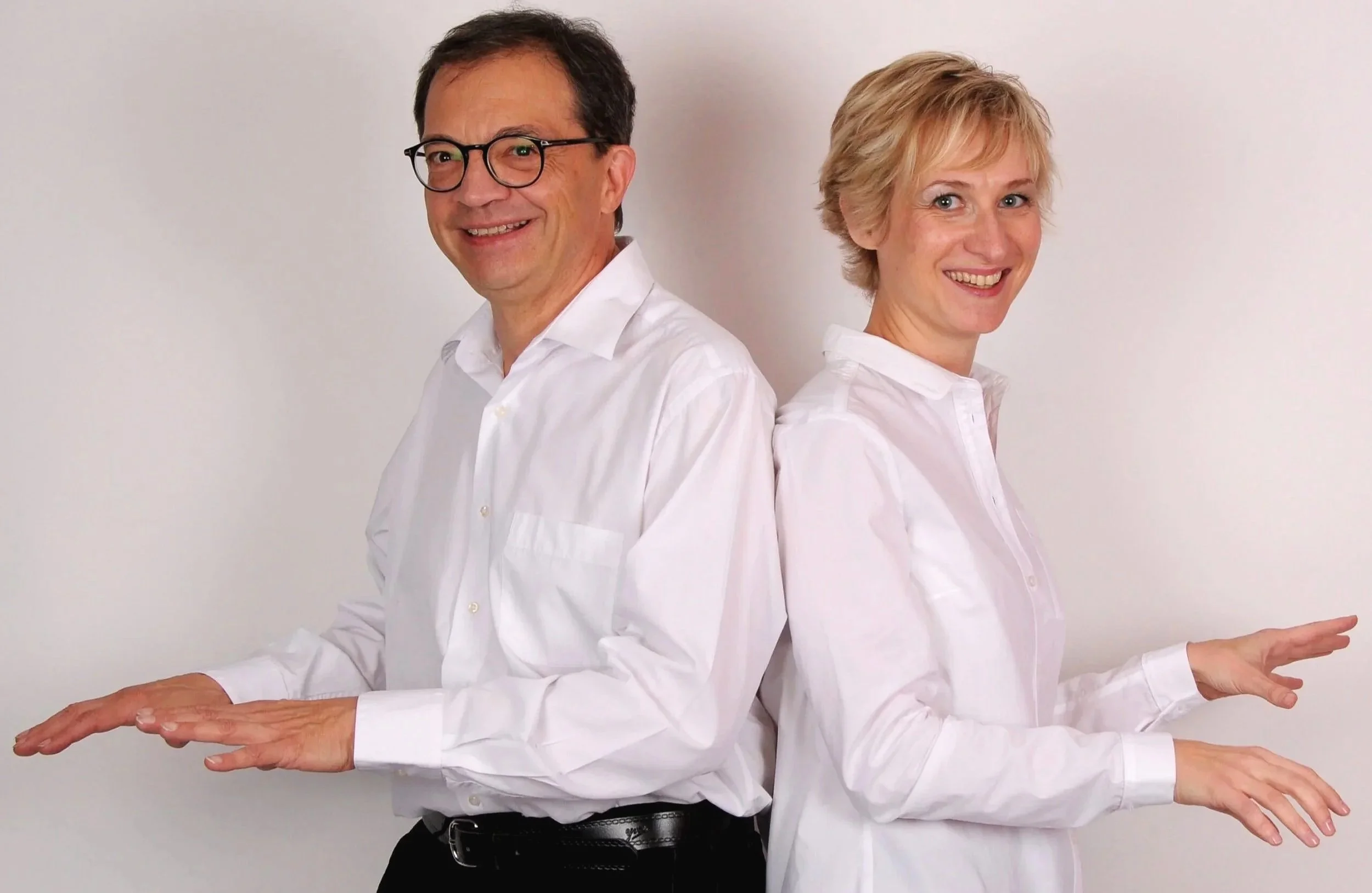 Un homme et une femme en chemises blanches, se tenant dos à dos, souriant, avec les bras tendus comme pour faire semblant de peser quelque chose.