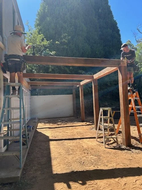 Two people working on building a wooden deck outdoors on a sunny day, with ladders and construction tools around.