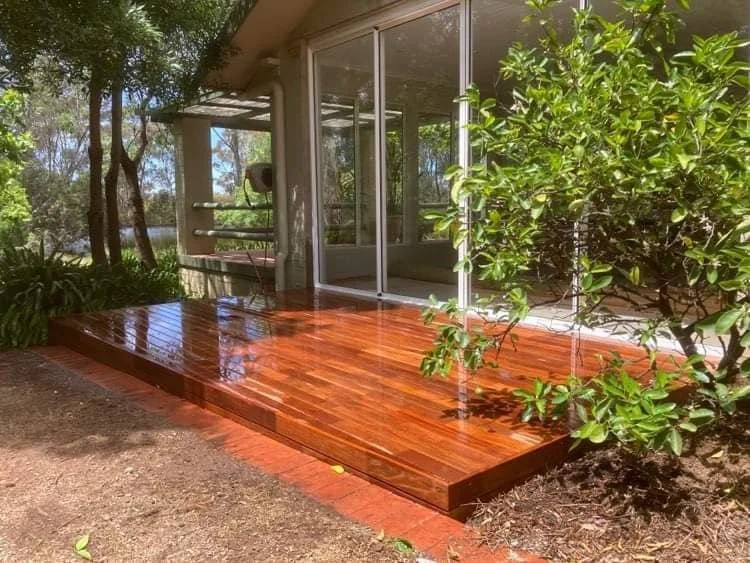 Wooden porch with shiny floor in front of a glass sliding door, surrounded by trees and bushes.