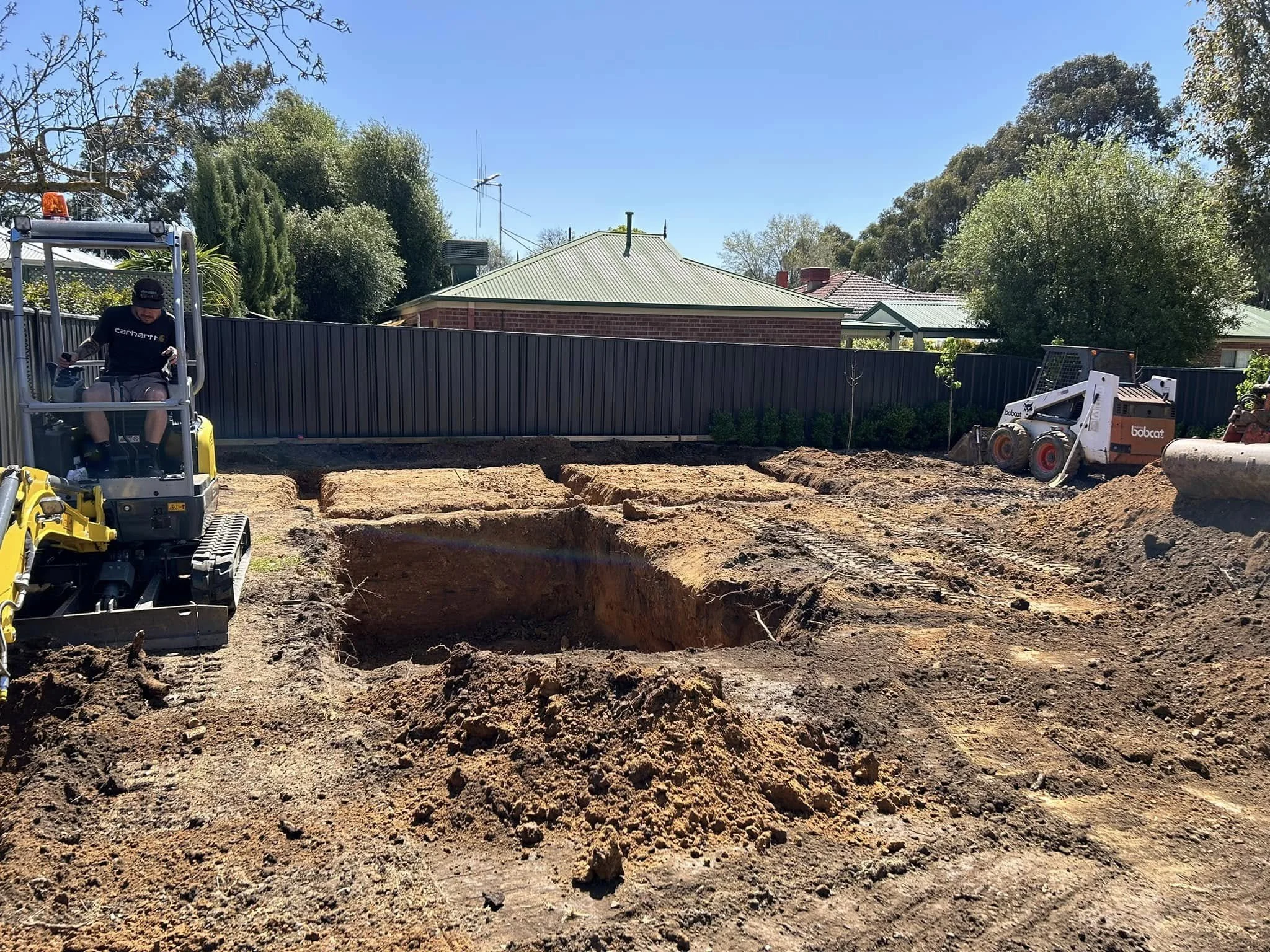 Construction site with two mini excavators digging in backyard of residential area, fences, houses, and trees in background.