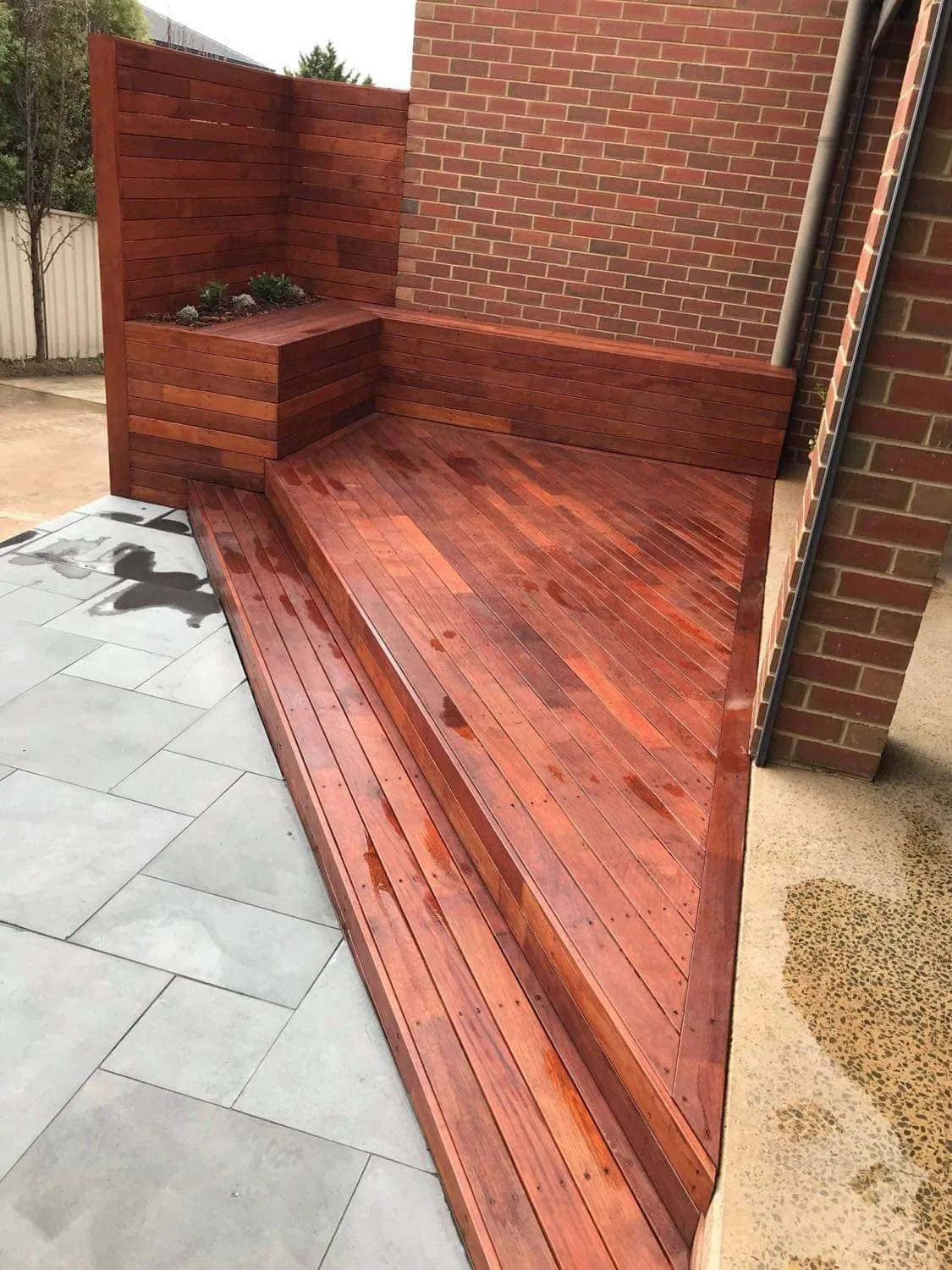 Wooden deck with curved edge adjacent to a brick wall, with some wet spots, and contrasting tile and concrete flooring nearby.
