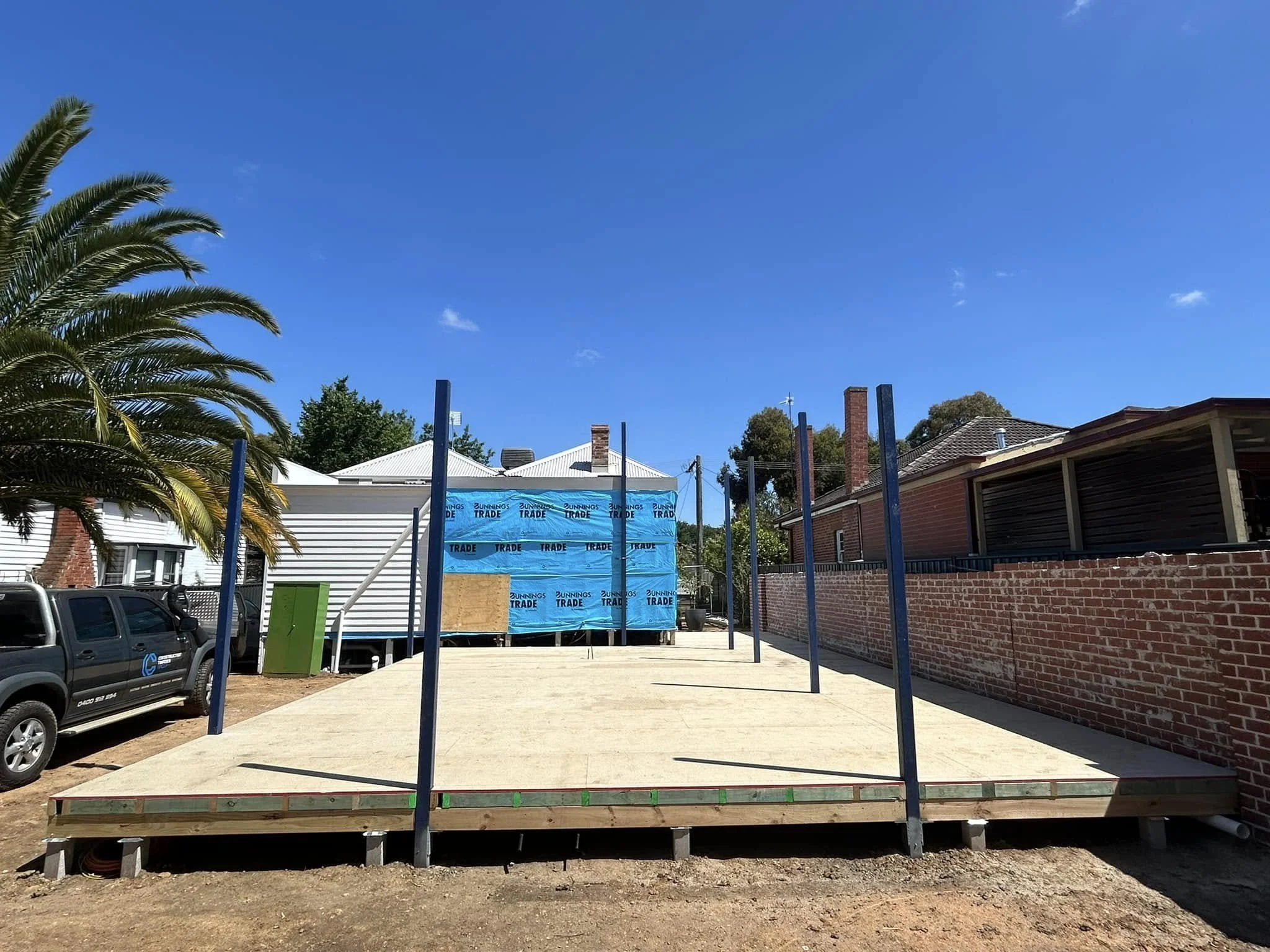 Construction site with a raised wooden platform, metal posts, a palm tree to the left, houses in the background, and a clear blue sky.