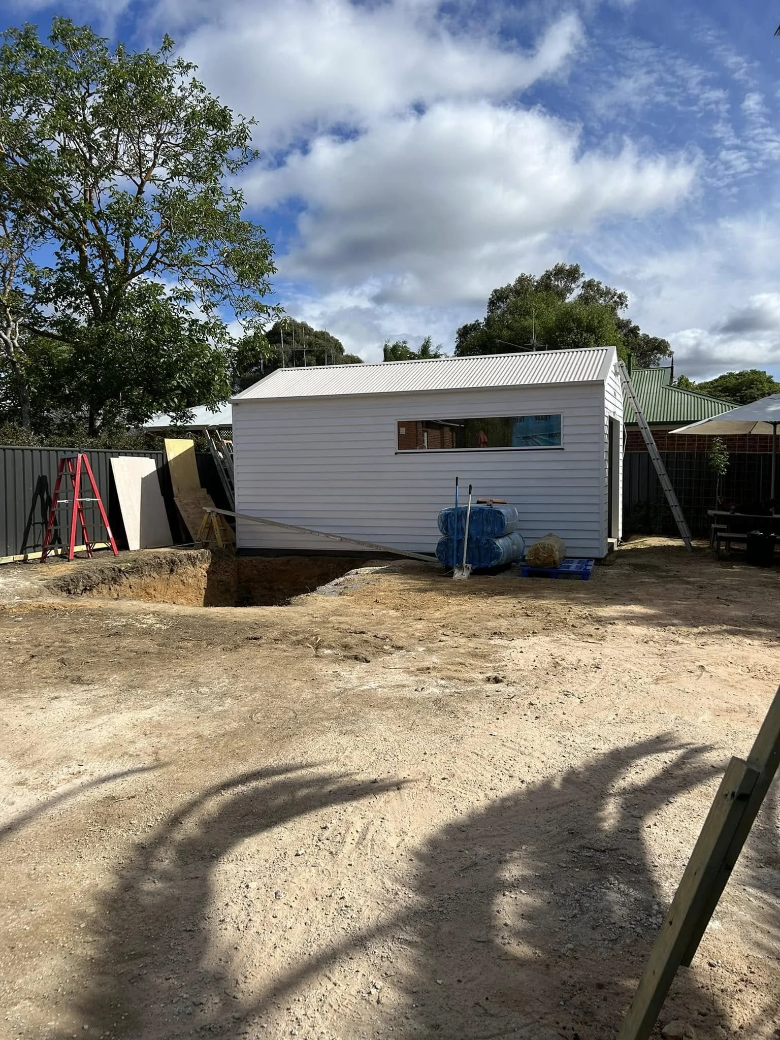 Construction site showing a small white building with a metal roof, surrounded by dirt and construction materials, with a partly cloudy sky overhead