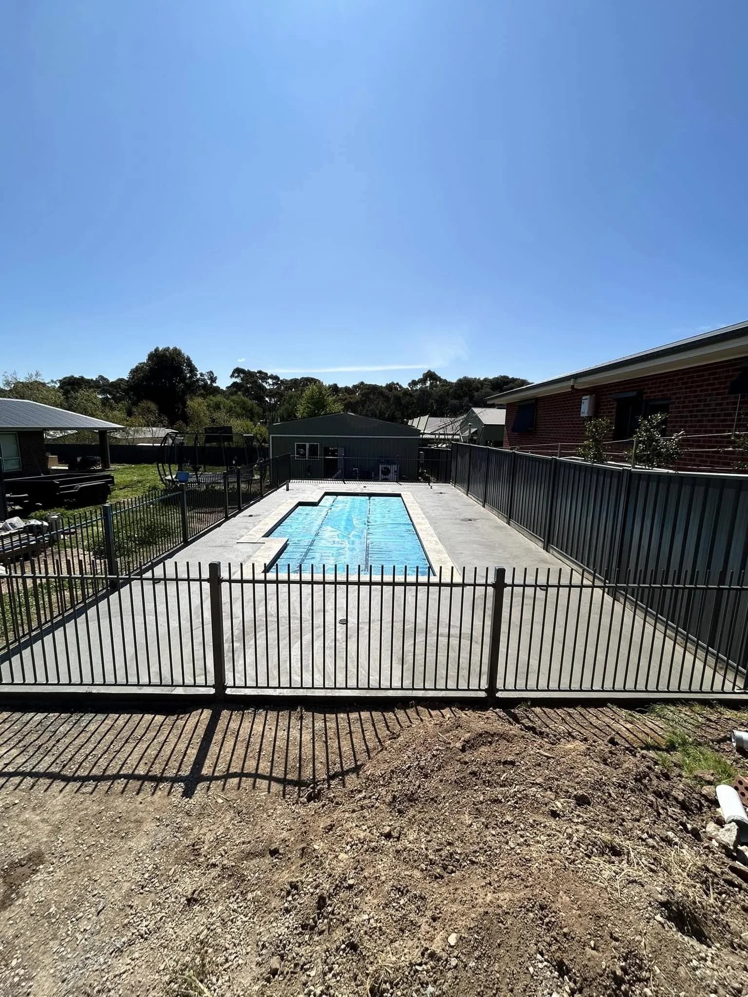 Backyard with a fenced-in swimming pool and concrete patio, neighboring houses, clear blue sky, and trees in the background.