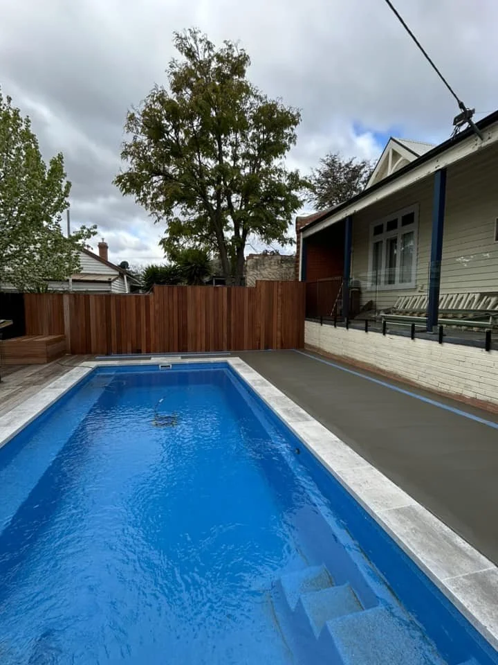 In-ground rectangular swimming pool with blue water in a backyard, enclosed by a wooden privacy fence, with a residential house towards the right and large trees in the background under a cloudy sky.