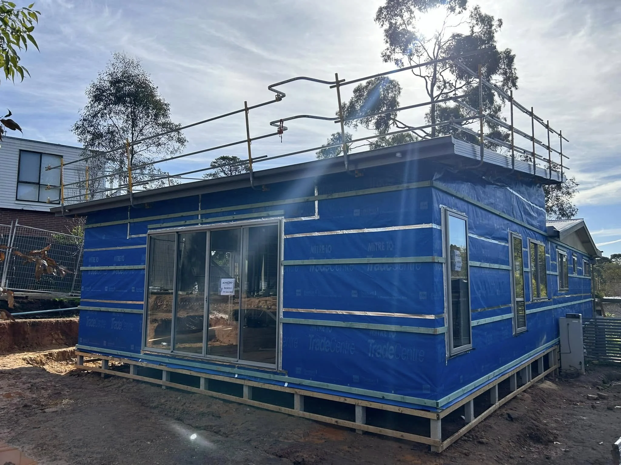 A house under construction wrapped in blue weather-resistant building material with windows and sliding glass doors installed, and a metal railing on the roof for safety.