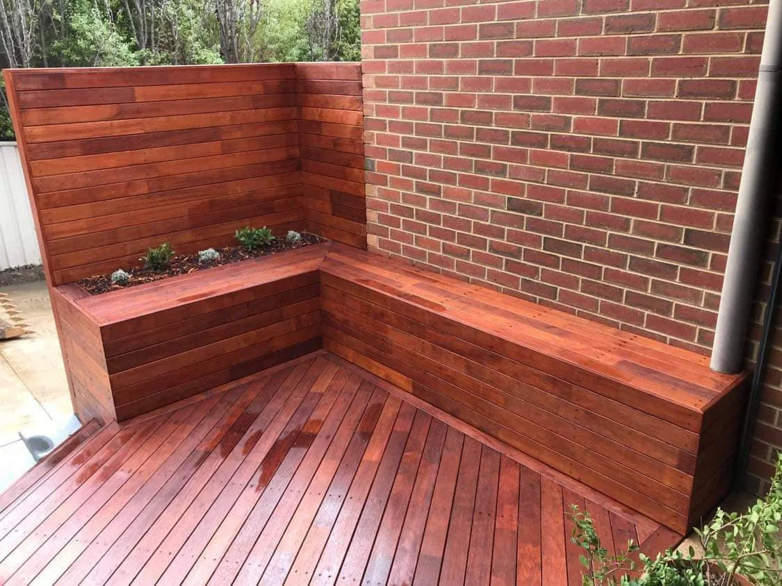 A corner outdoor wooden bench with planters against a brick wall, on a newly stained wooden deck.