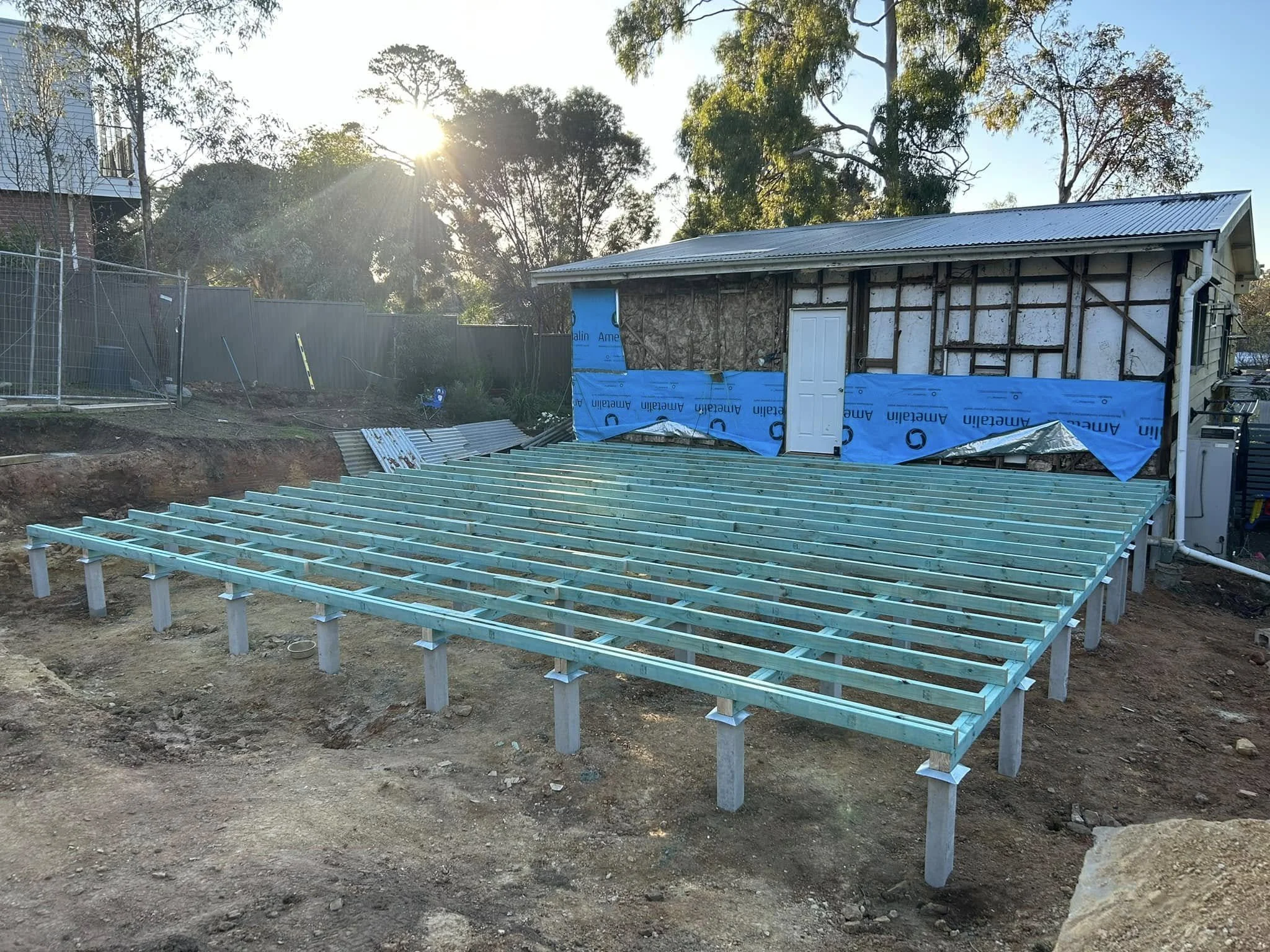 Wooden framework under construction on a dirt yard, with a small building in the background partly covered in blue weatherproofing material and a shed, trees, and the sun setting behind them.
