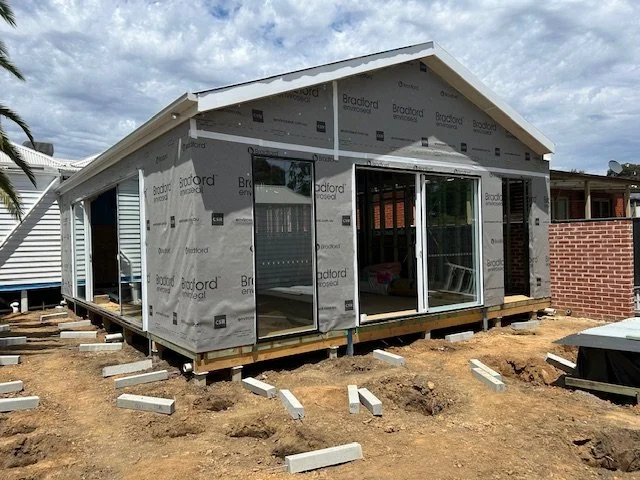Under construction extension partly covered in blue weatherproofing materialon a dirt yard, with a small building in the background.
