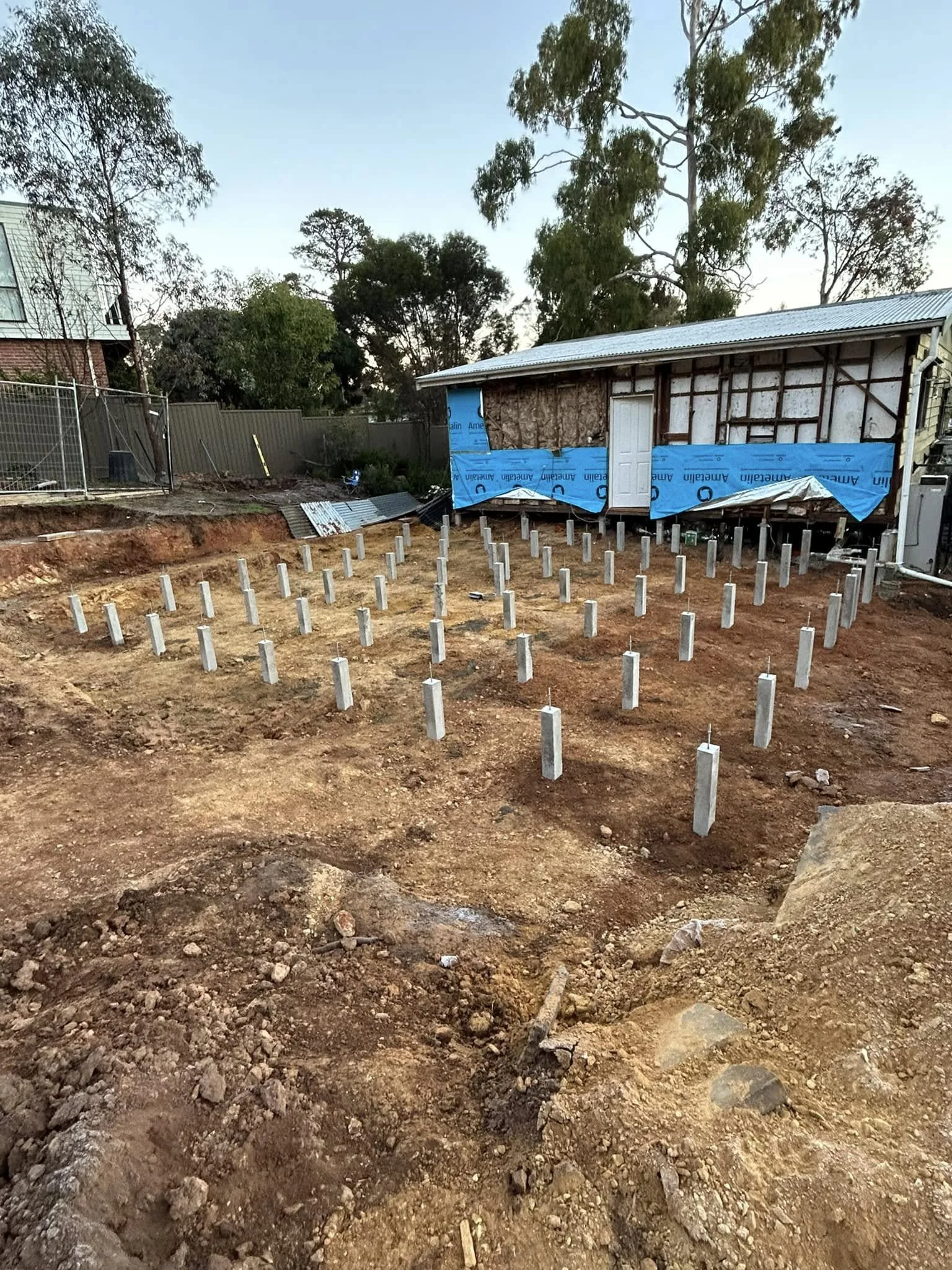 Construction site with numerous concrete footing piers installed, and a partially built shed in the background.