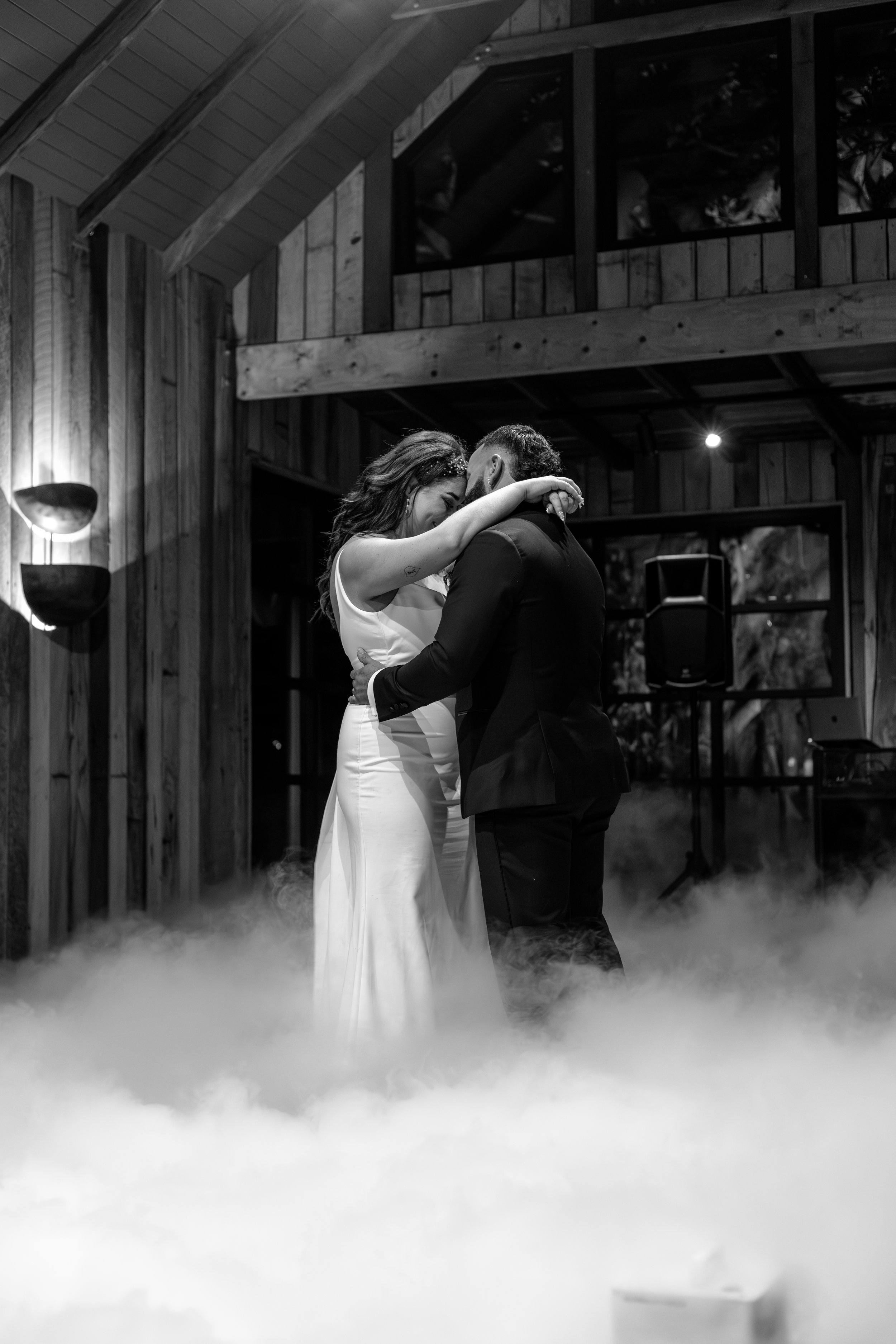 A couple dancing closely at their wedding reception, with mist on the floor and wooden interior in the background.