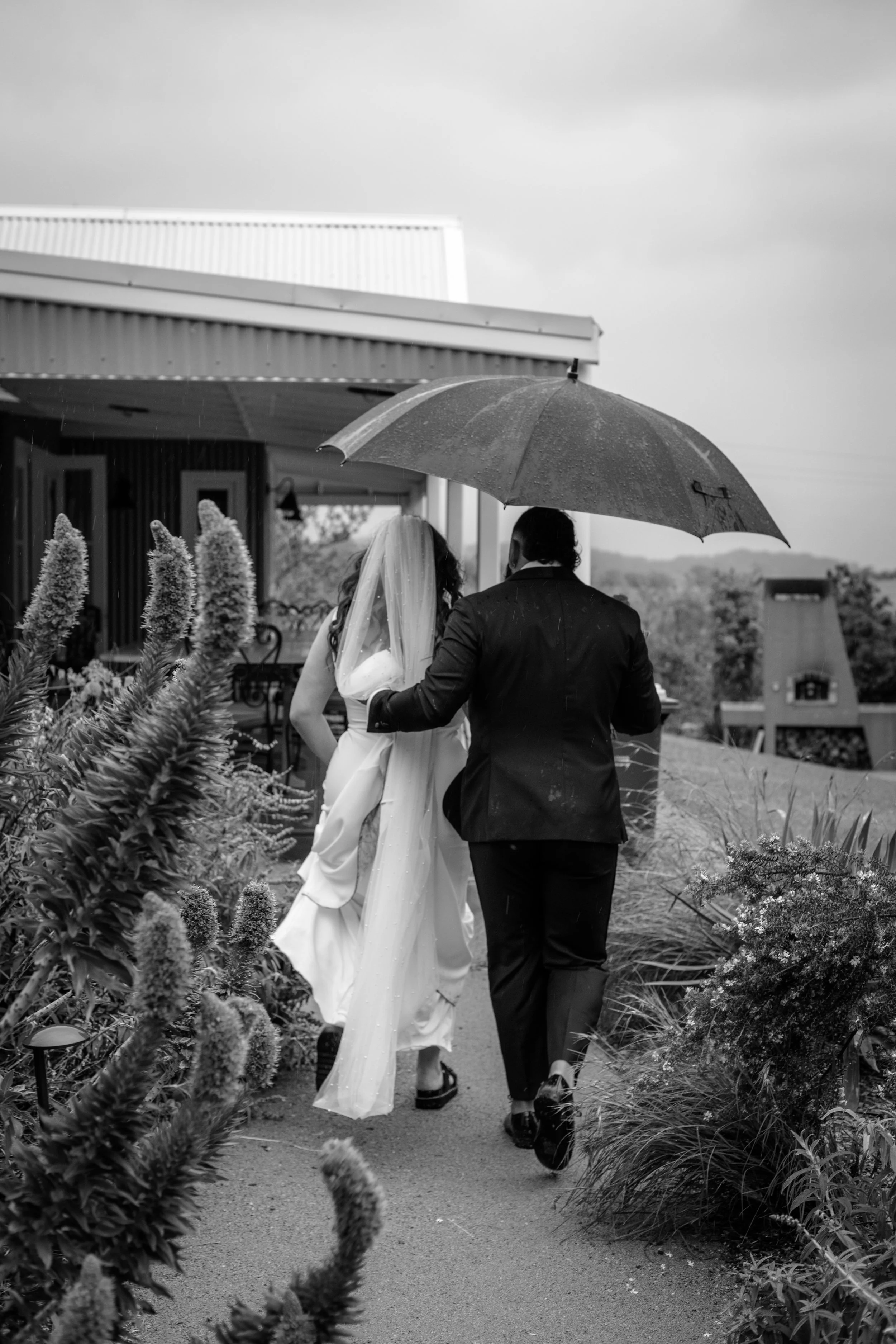 A couple, possibly bride and groom, walking under an umbrella on a rainy day outdoors.