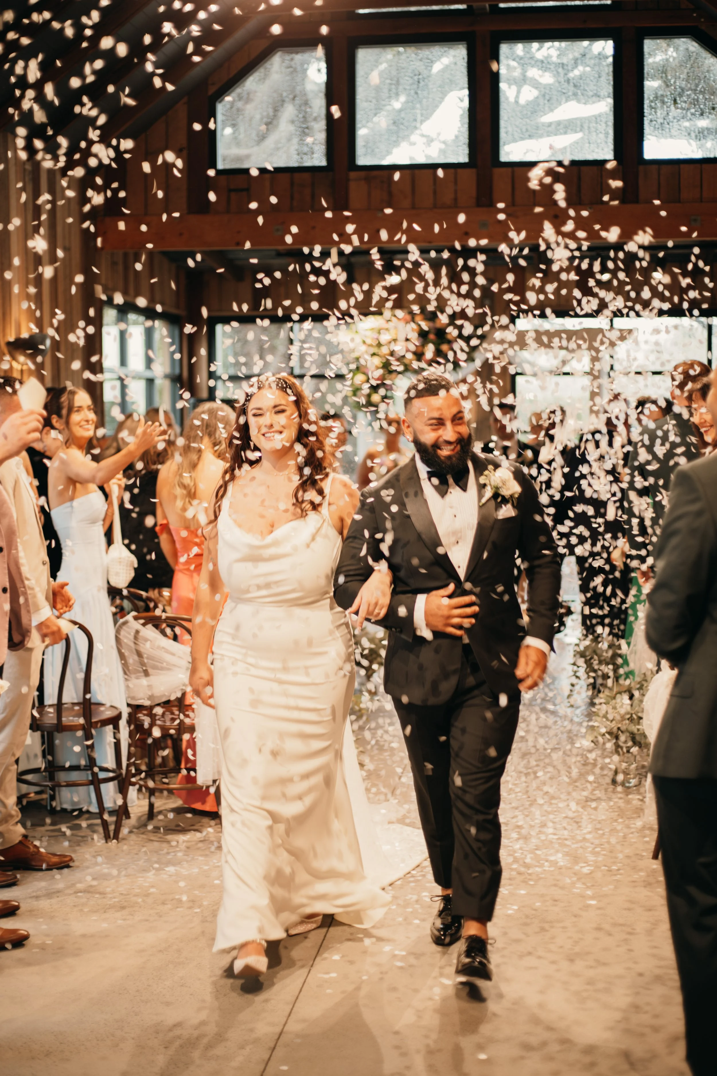 A newlywed couple walks down the aisle surrounded by friends and family as white flower petals are thrown in the air, celebrating their wedding in a warmly lit indoor venue with large windows and wooden interior.