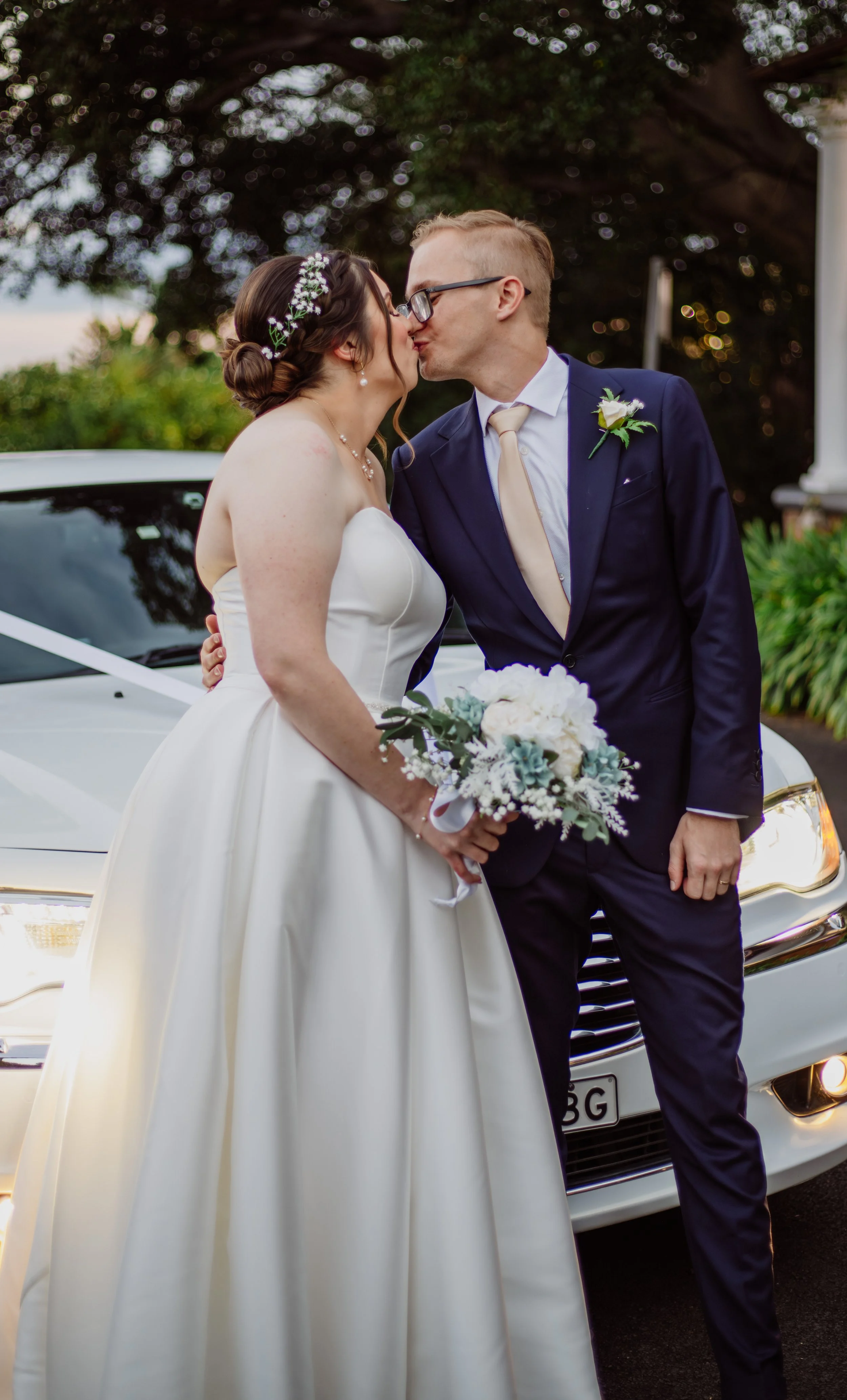 A bride and groom sharing a kiss in front of a white luxury car with a black grille, during a wedding celebration outdoors, with trees and a house in the background.