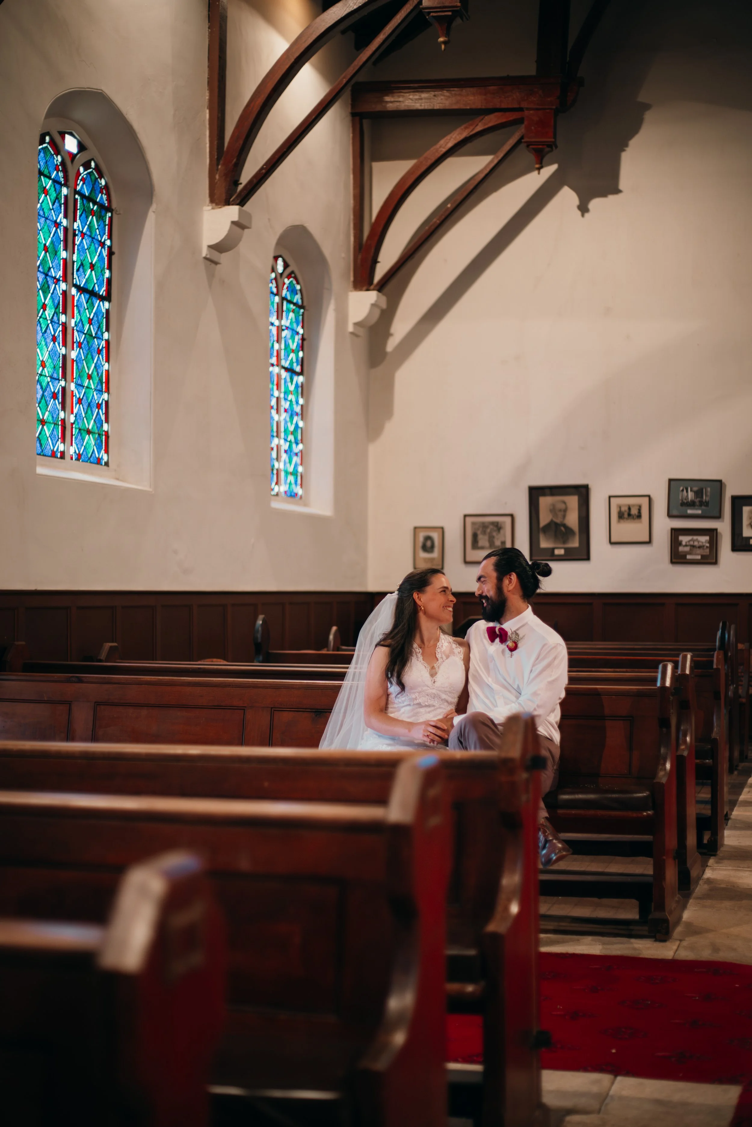 A bride and groom sitting together inside a church, smiling at each other, with stained glass windows and framed photographs on the wall behind them.