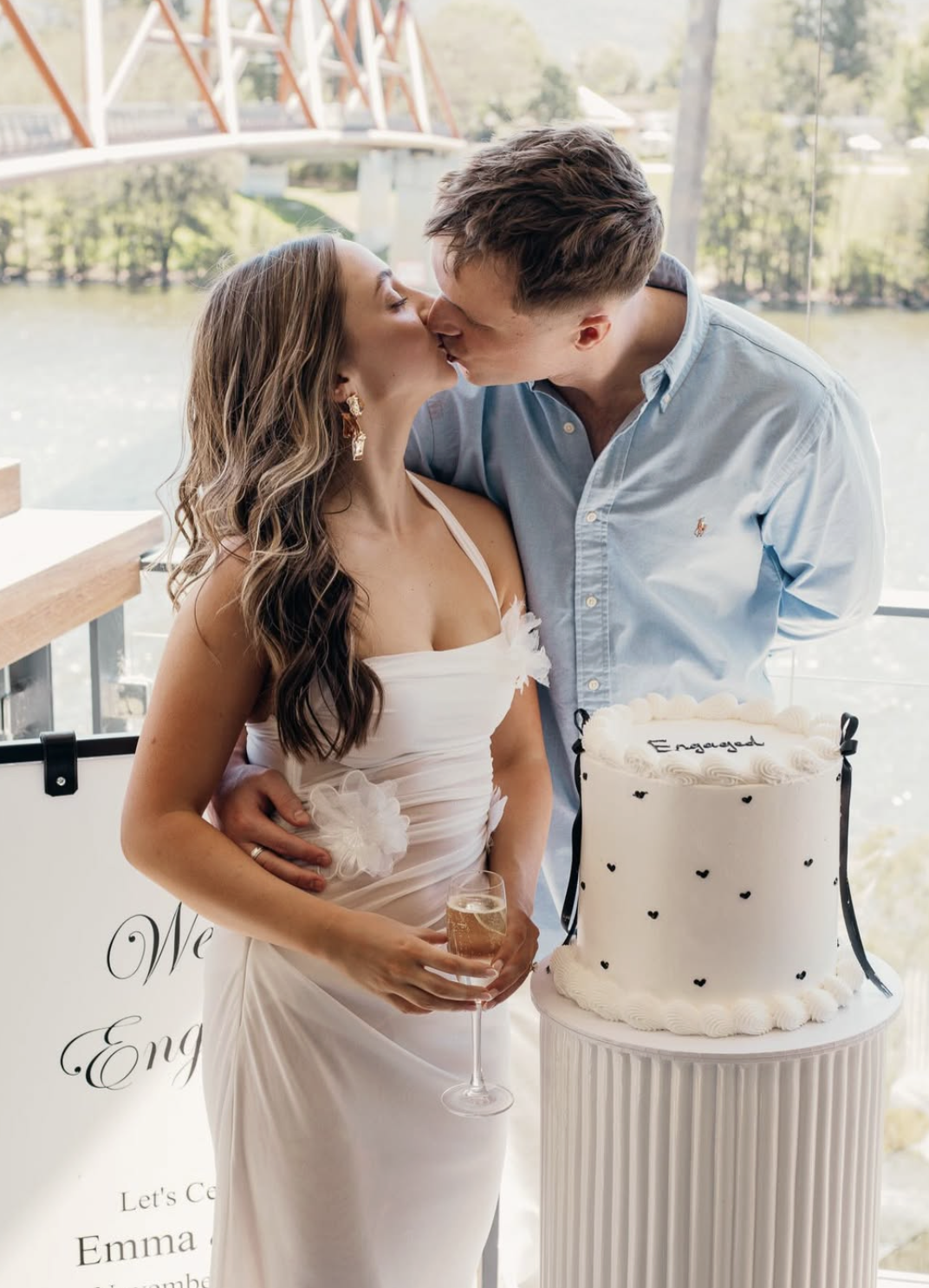 A couple is kissing at their wedding reception. The bride is holding a glass of champagne, and a wedding cake with "Engaged" written on top is beside them. They are standing near a large window overlooking a river and a bridge.