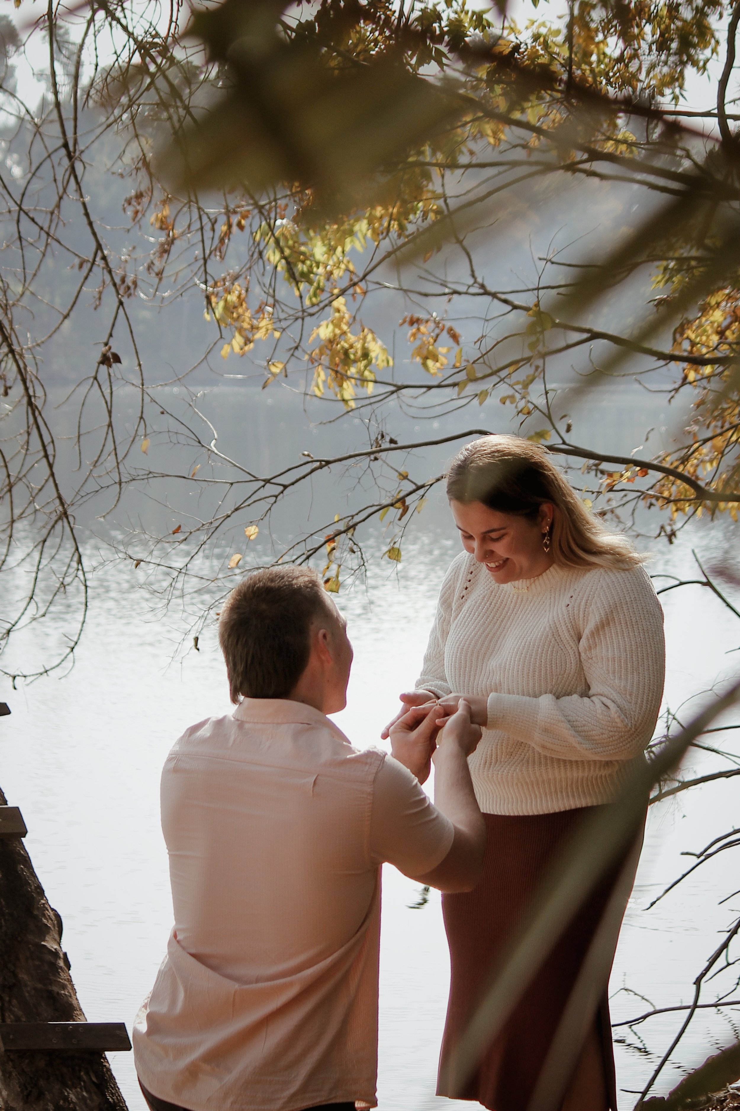 A man proposing marriage to a woman by a lake surrounded by trees, with golden leaves and sunlight filtering through the branches.