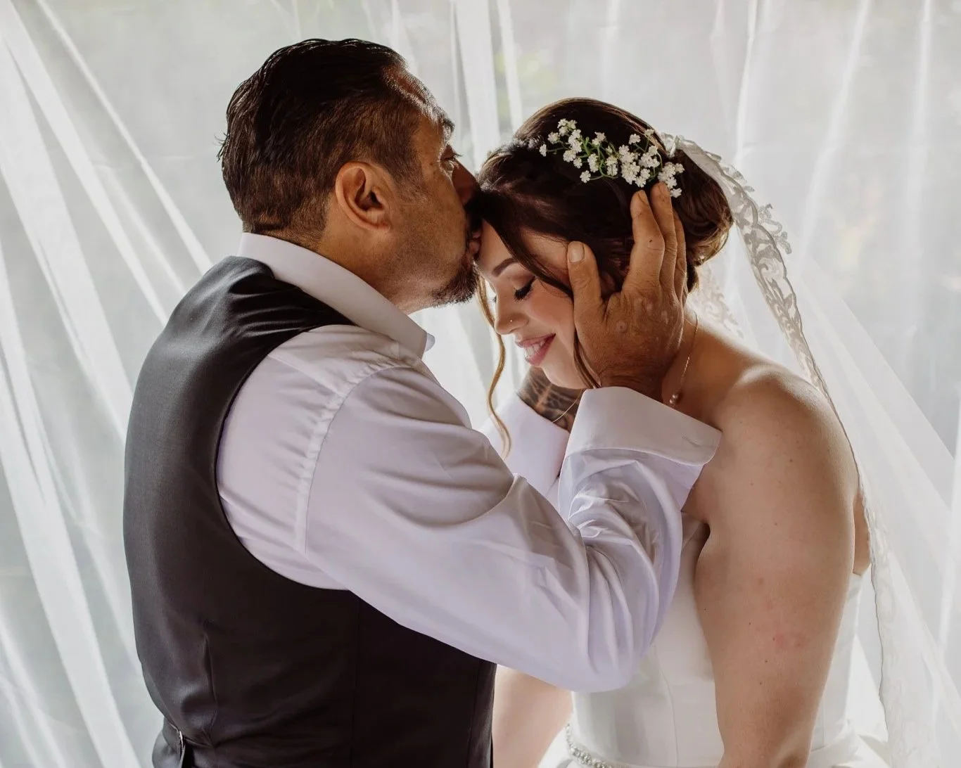 A groom gently kissing a bride on her forehead during their wedding, with both sharing a tender moment.