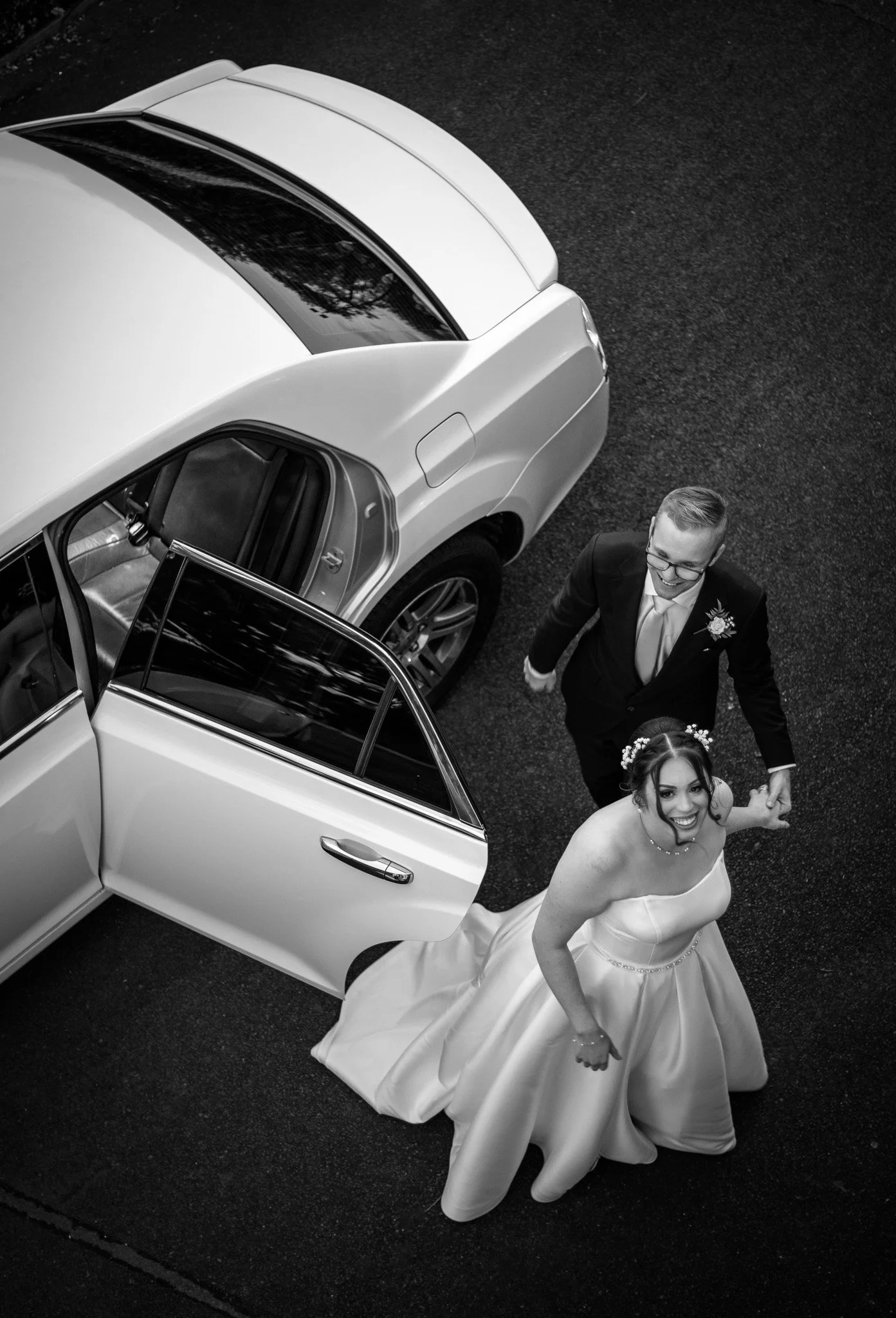 Black and white photo of a smiling bride and groom holding hands, standing next to a white luxury car with an open door, on a dark asphalt road.