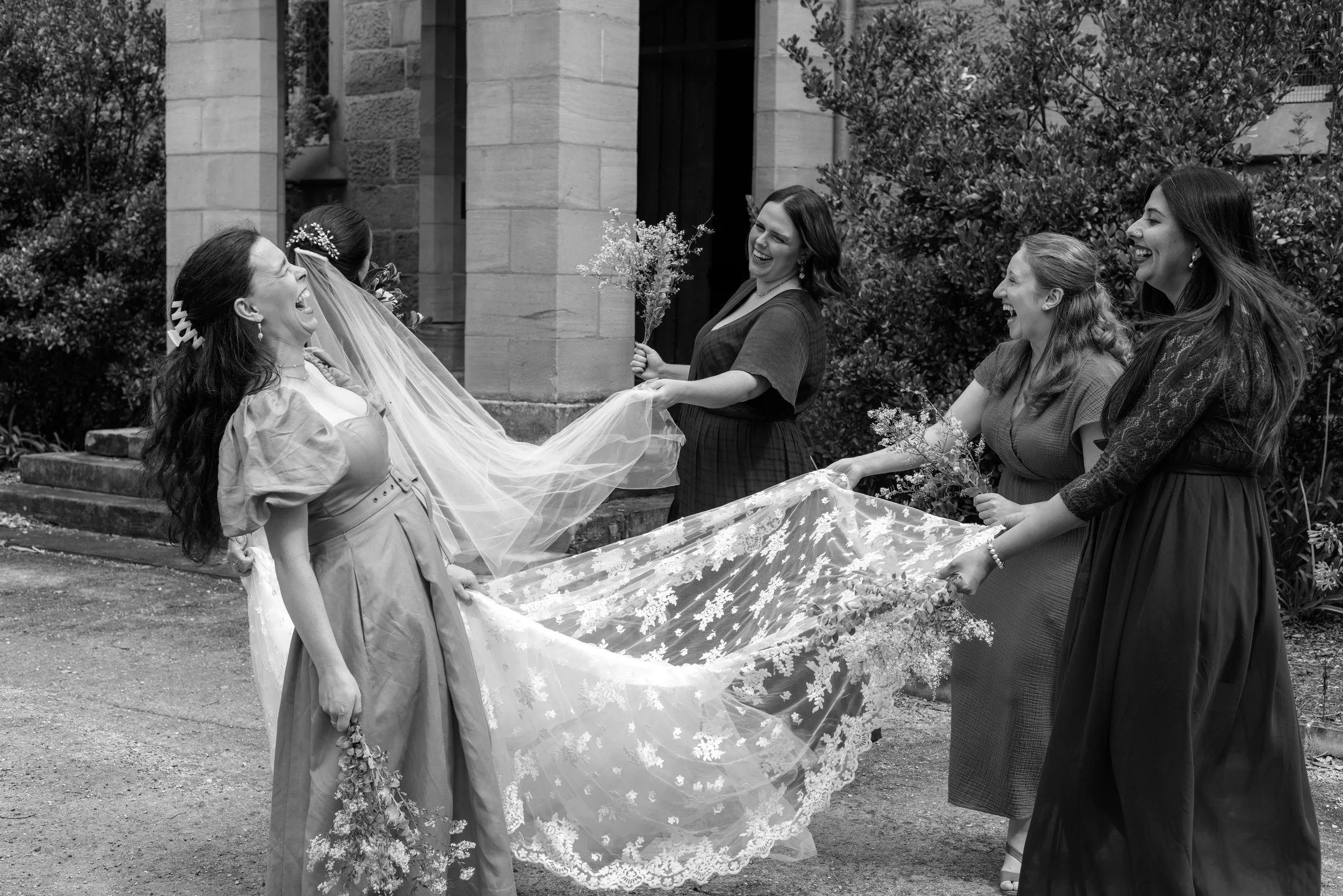 A group of women dressed as bridesmaids holding the train of a bride's wedding gown outside near a stone building and foliage, all laughing and smiling.