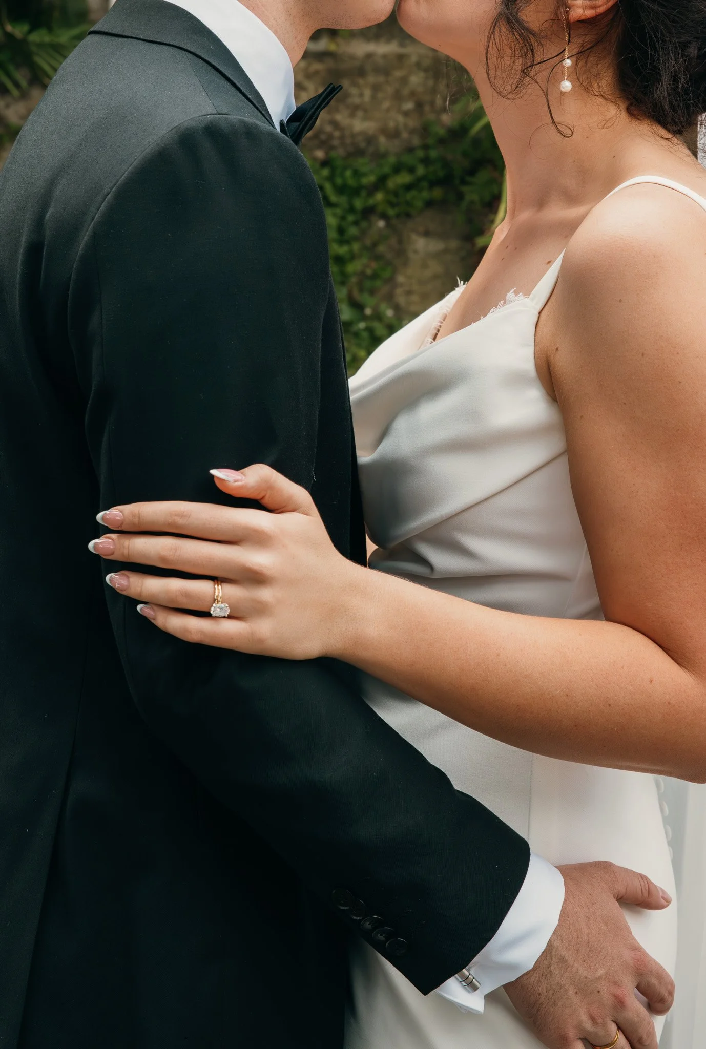 Close-up of a bride and groom on their wedding day, showing the bride's left hand with an engagement ring and wedding band, resting on the groom's chest, with the groom dressed in a black tuxedo and the bride in a white wedding gown.
