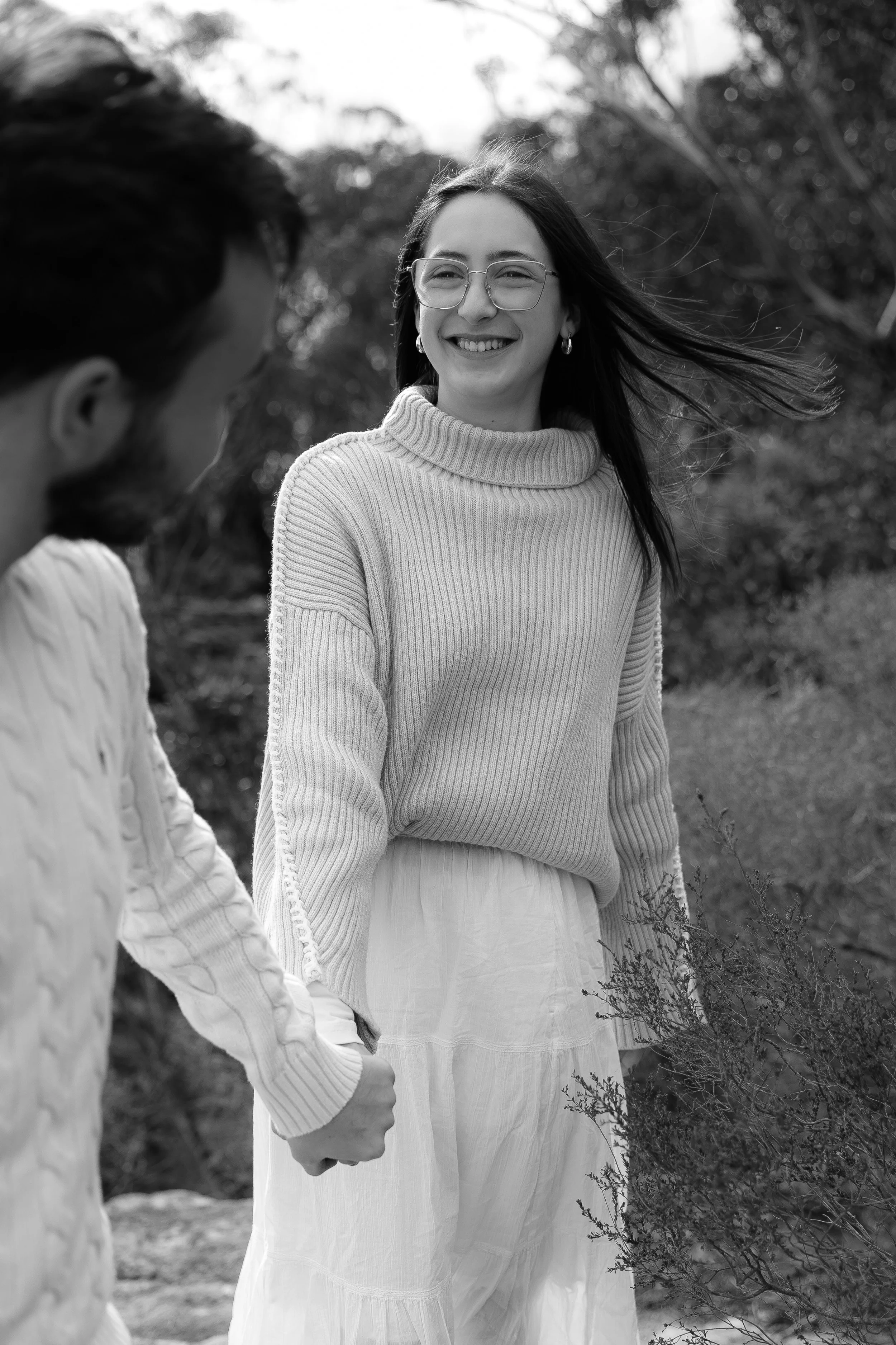 A woman with glasses and long hair smiling while holding hands with a man, outdoors with trees in the background, in black and white.