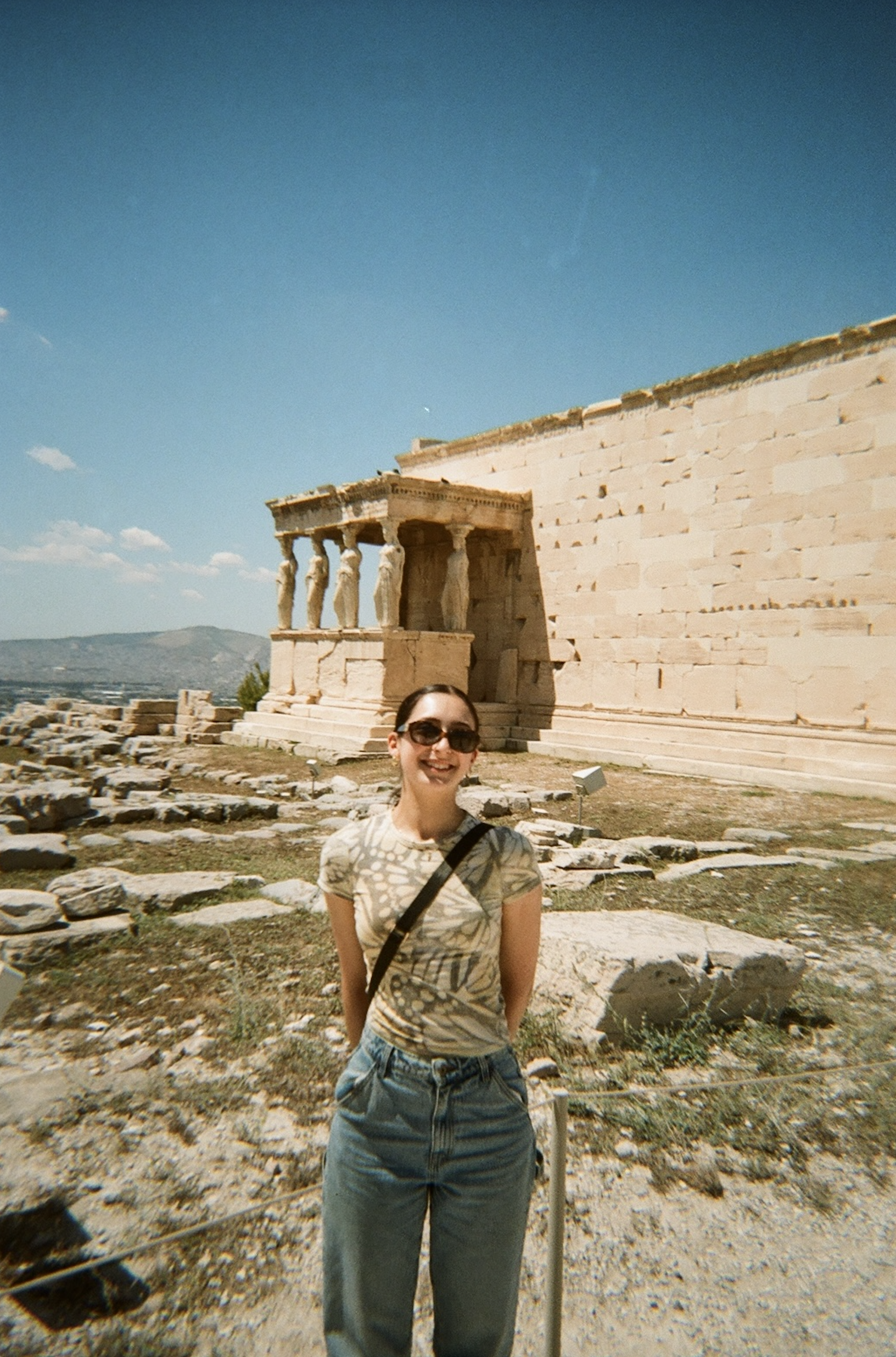 A woman posing outdoors in front of ancient Greek ruins, wearing sunglasses, a patterned t-shirt, and casual pants.