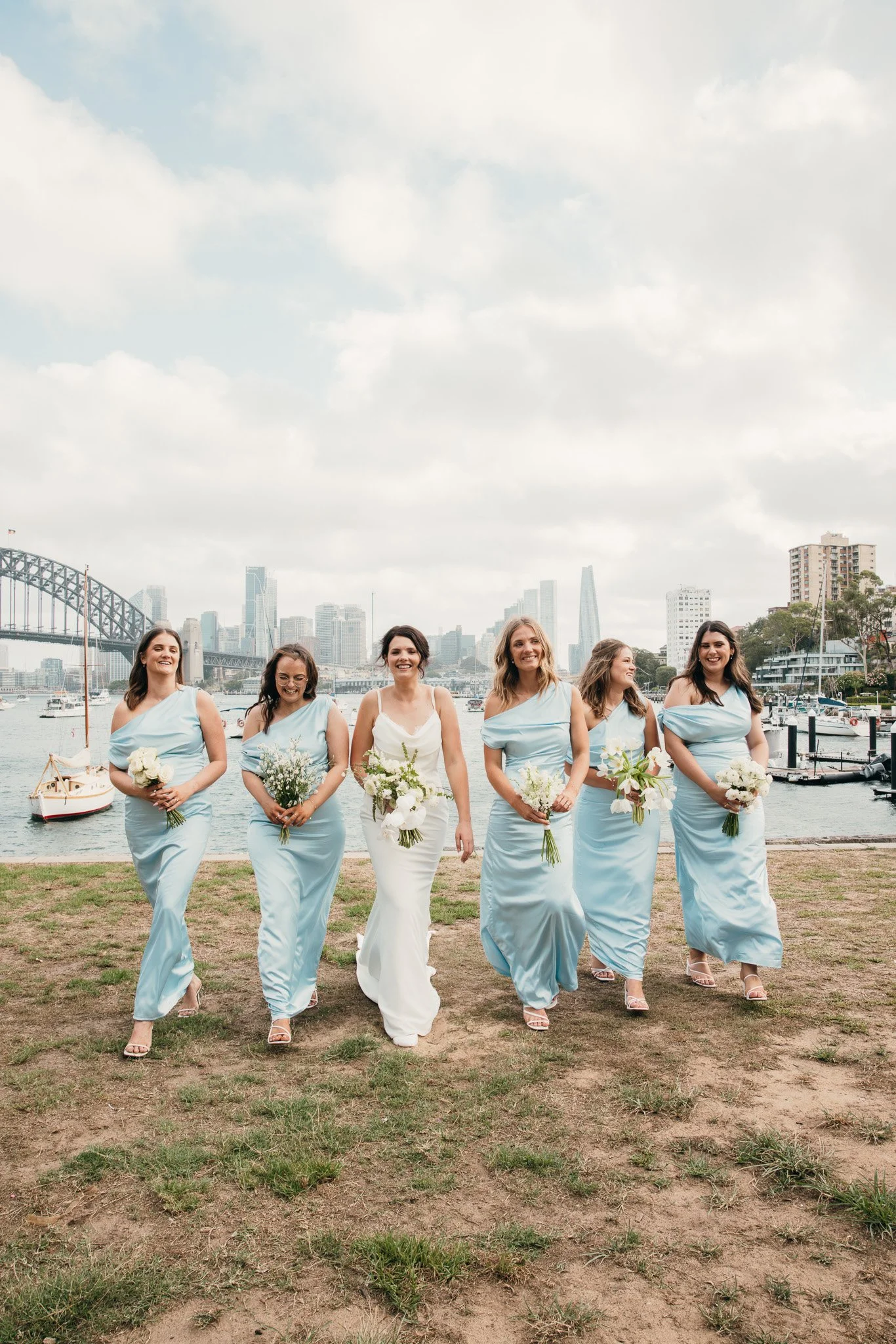 Six women in bridesmaid dresses walking on grass near a waterway with boats, city skyline, and bridge in the background.
