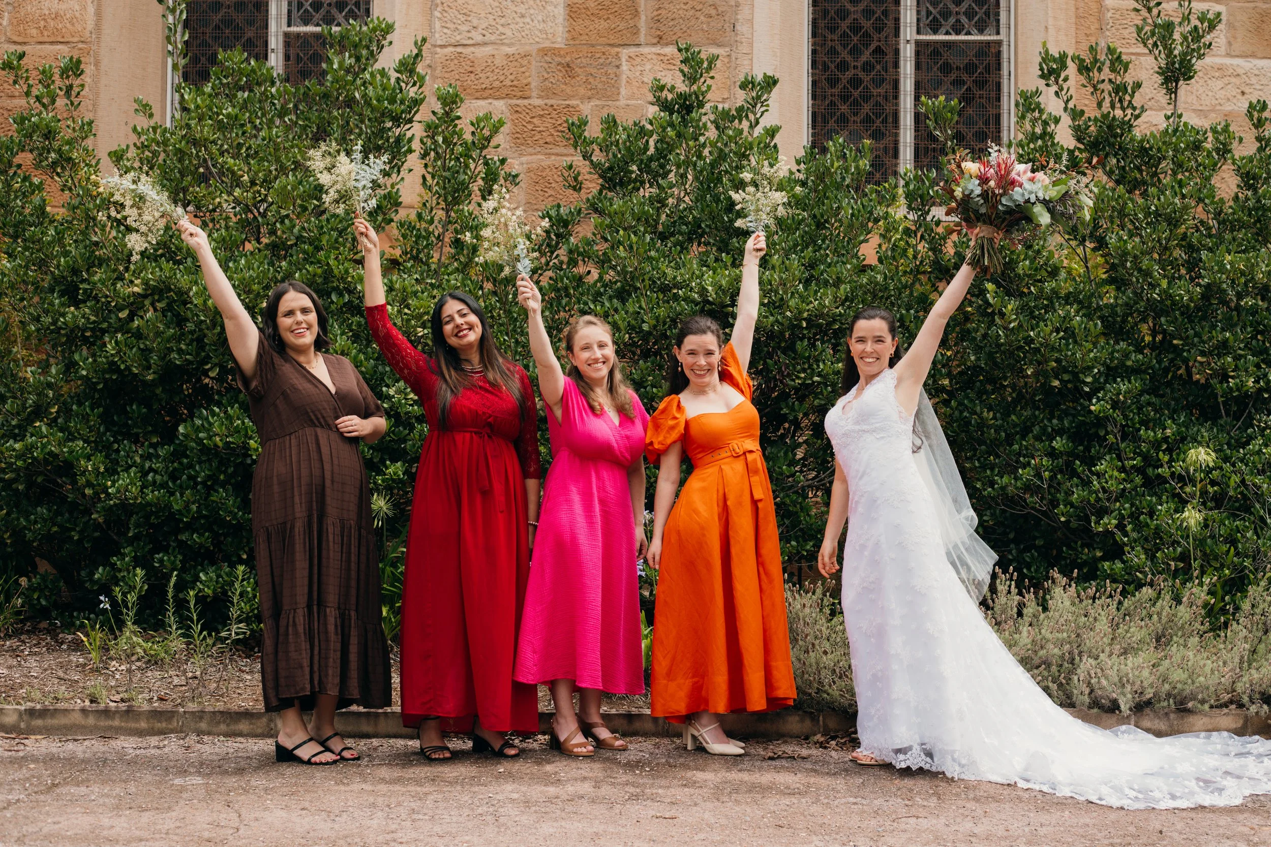 Five women celebrating outdoors, with the bride in a white wedding dress and four women in colorful dresses. The women are smiling, raising bouquets, and standing in front of green bushes and a stone building.