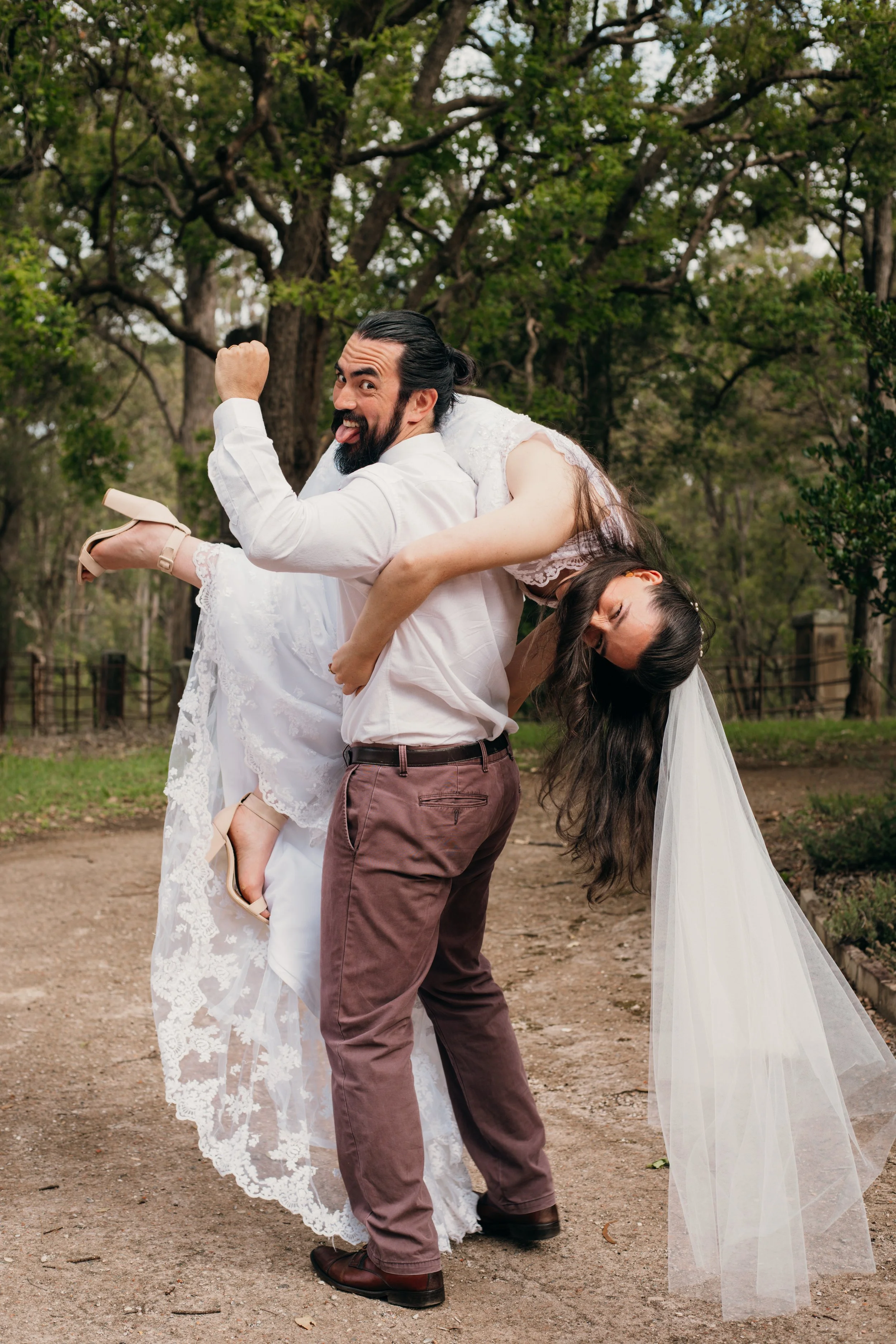A joyful couple dressed in wedding attire enjoying a playful moment outdoors in a wooded area. The man is lifting the woman and showing his muscles while she leans back with her eyes closed.