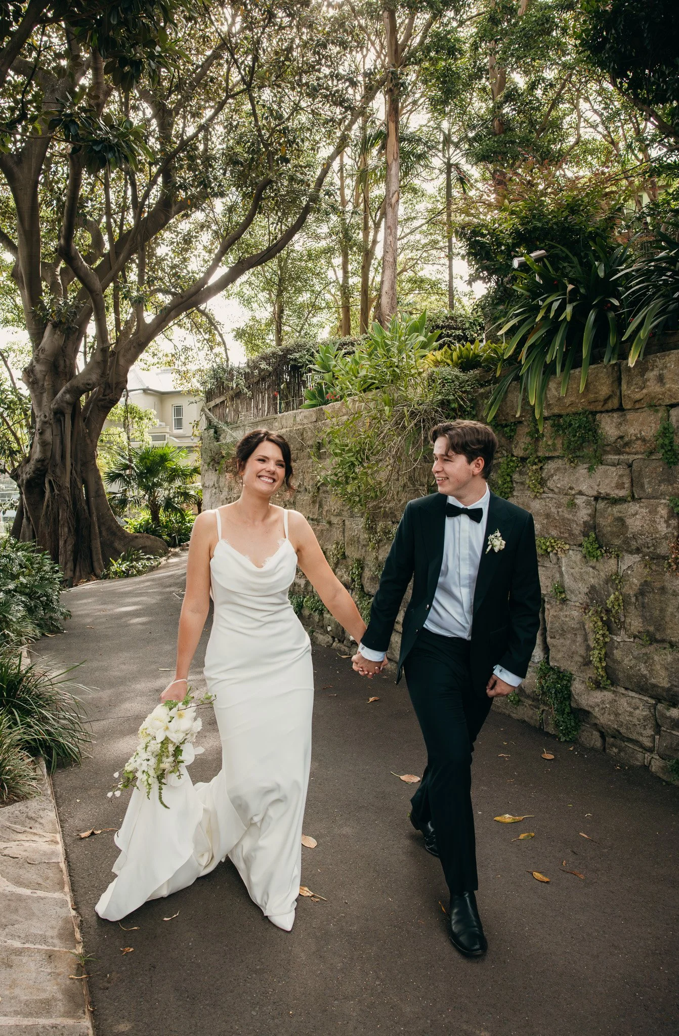 A bride and groom holding hands and walking outdoors in a lush garden with trees and stone walls, dressed in wedding attire and smiling.