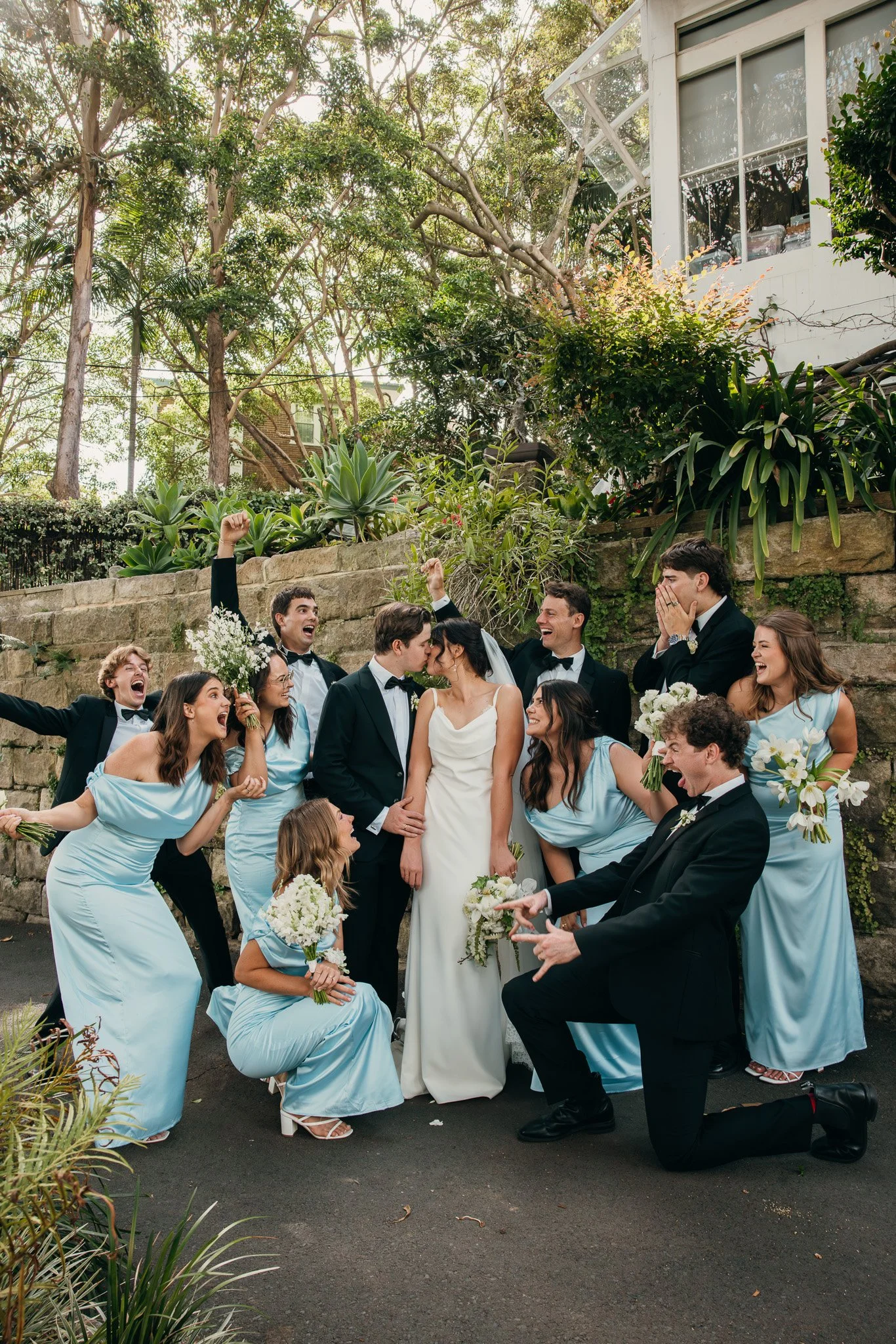 A wedding celebration with the bride, groom, and wedding party outdoors. The group is smiling, laughing, and celebrating, with some members holding flowers. The bride and groom are in the center, sharing a moment, surrounded by friends and family.