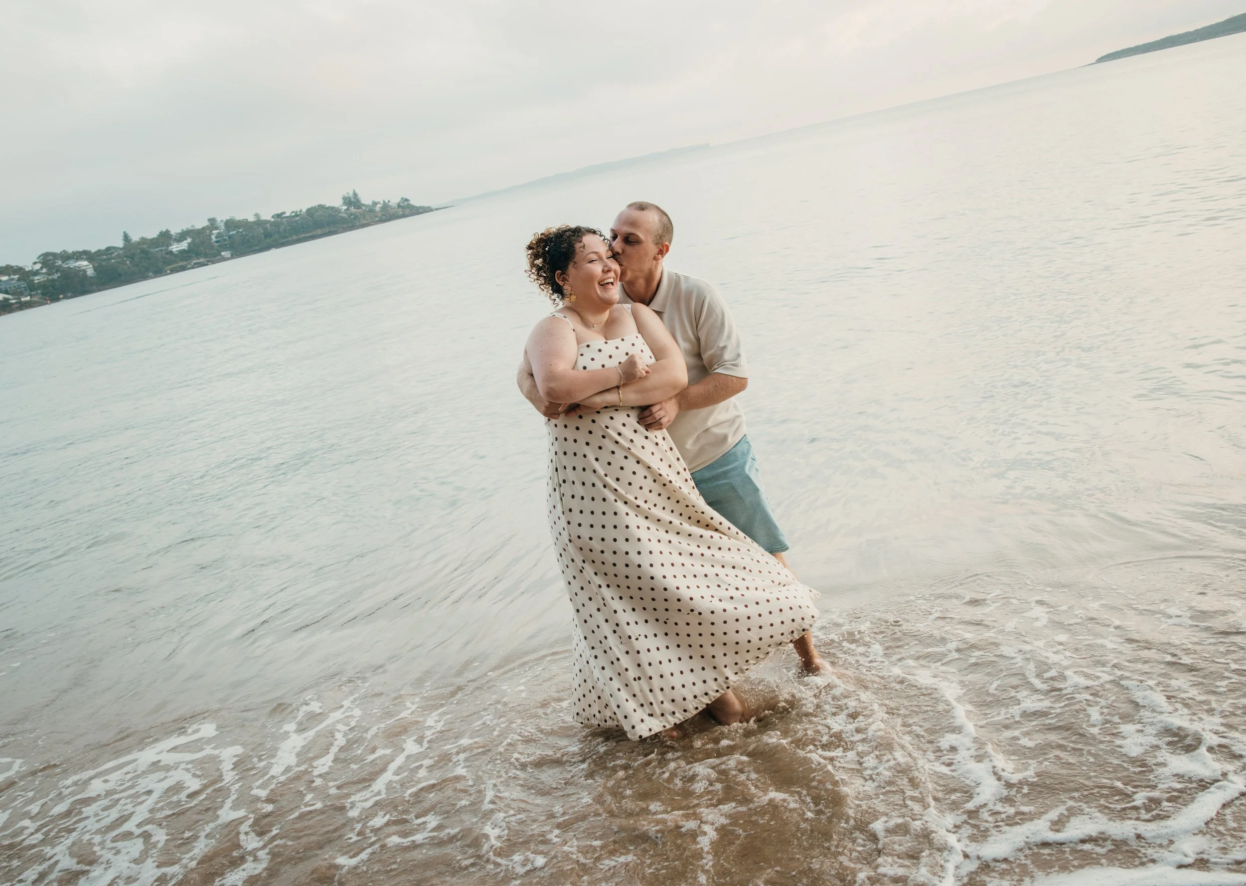A couple hugging and laughing in the water at the beach, with a scenic shoreline and cloudy sky in the background.