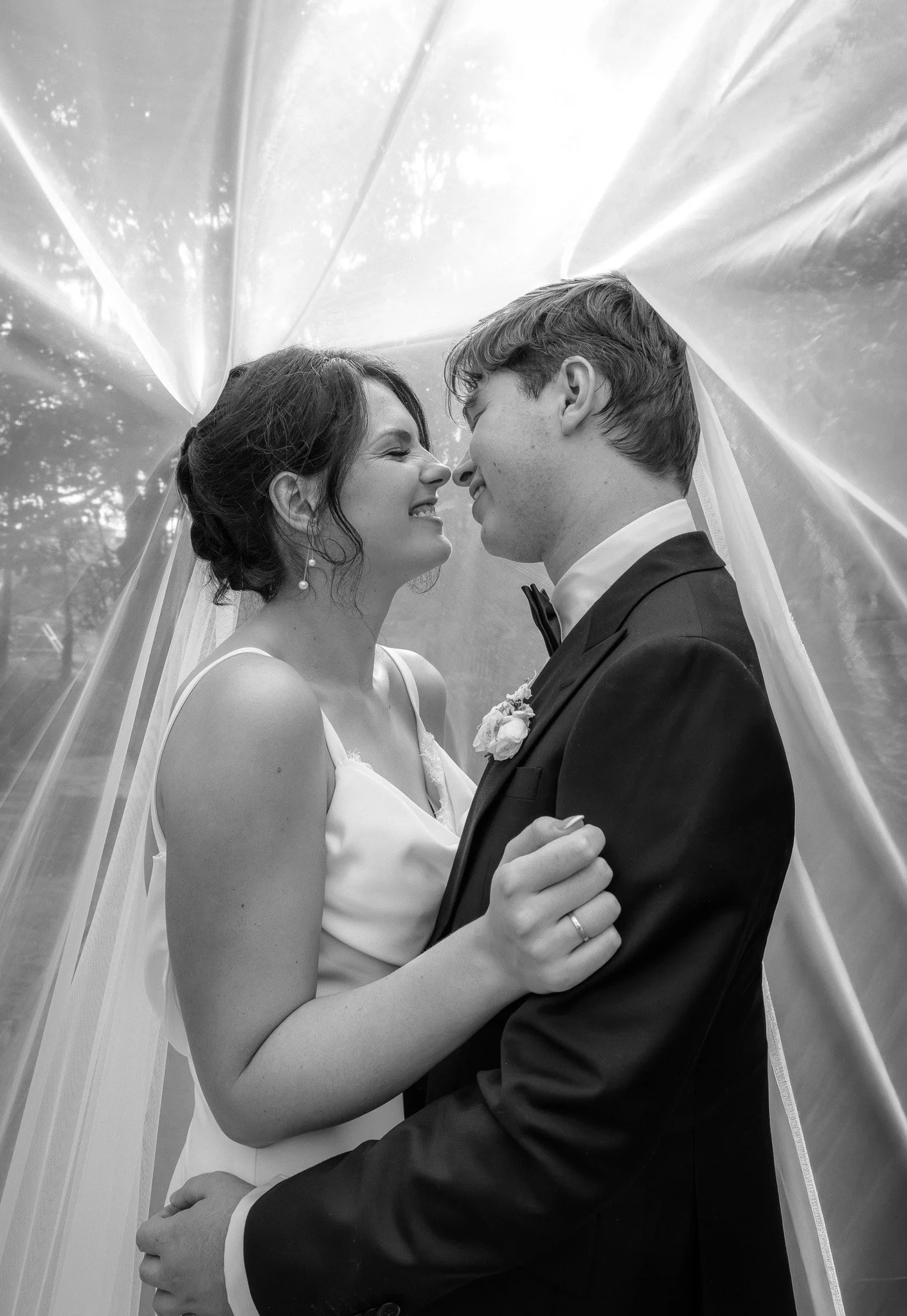 Black and white photo of a bride and groom smiling nose to nose under a transparent veil, outdoors with trees in the background.