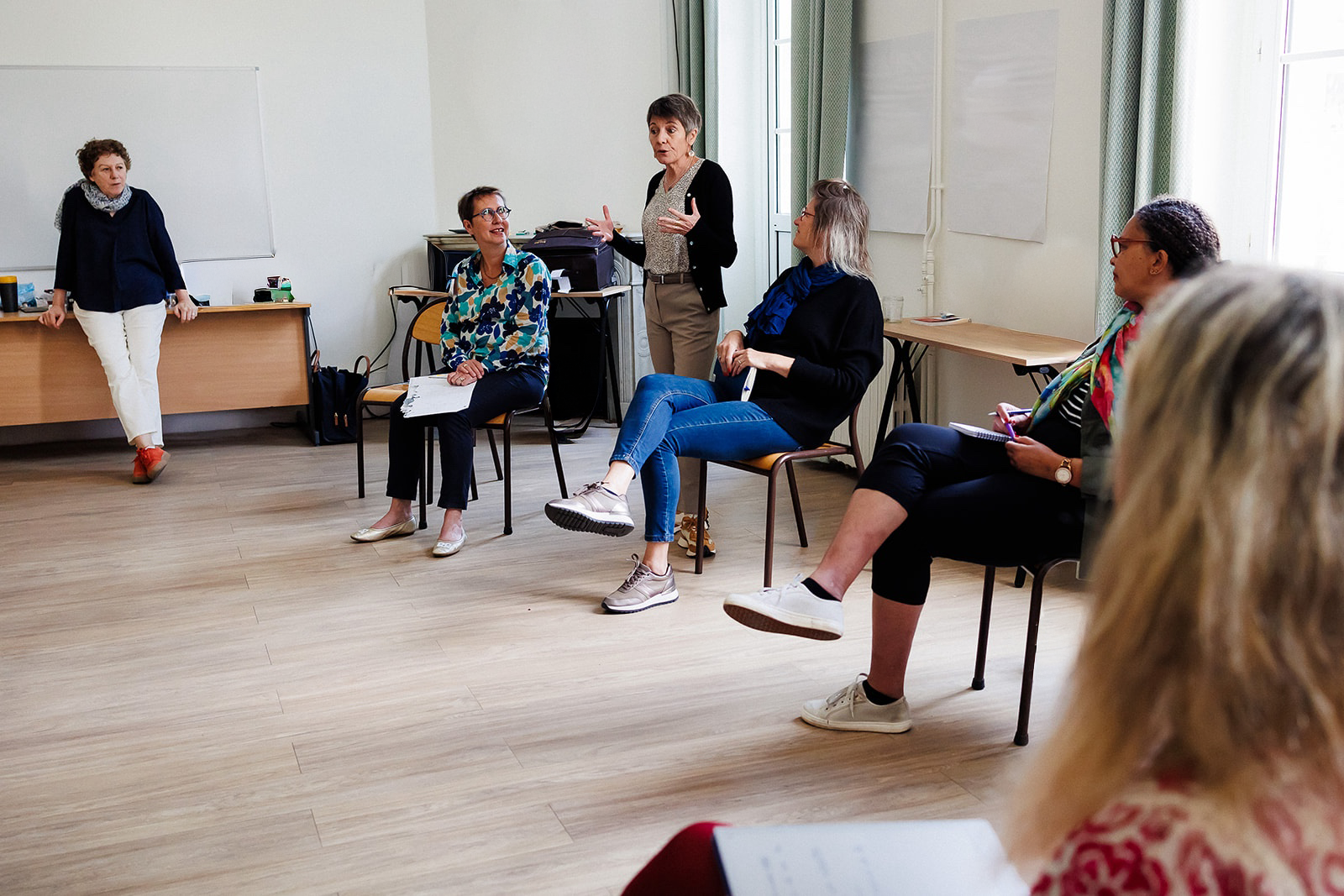 Un groupe de femmes assises dans une salle de réunion, écoutant une femme debout qui semble parler ou faire une présentation.