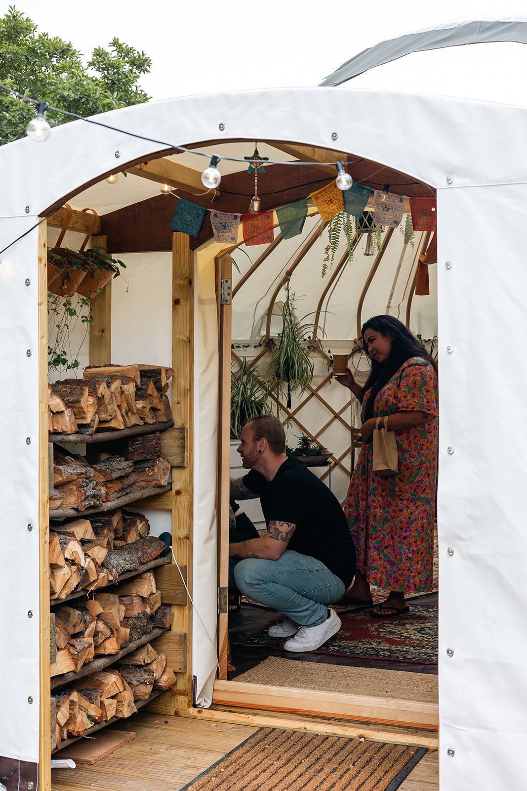A man and woman inside a cozy backyard wood storage tent, with wood logs stacked on shelves, string lights, and hanging plants, enjoying a warm drink.