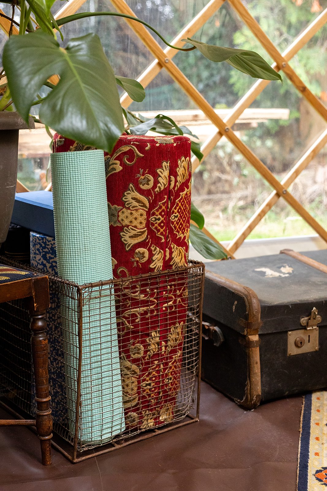 A wire basket containing a rolled blue yoga mat, a rolled green textured yoga mat, and a decorative red and gold embroidered fabric. The basket is next to a dark wooden table and a black vintage suitcase, with a potted plant on the left and a wooden