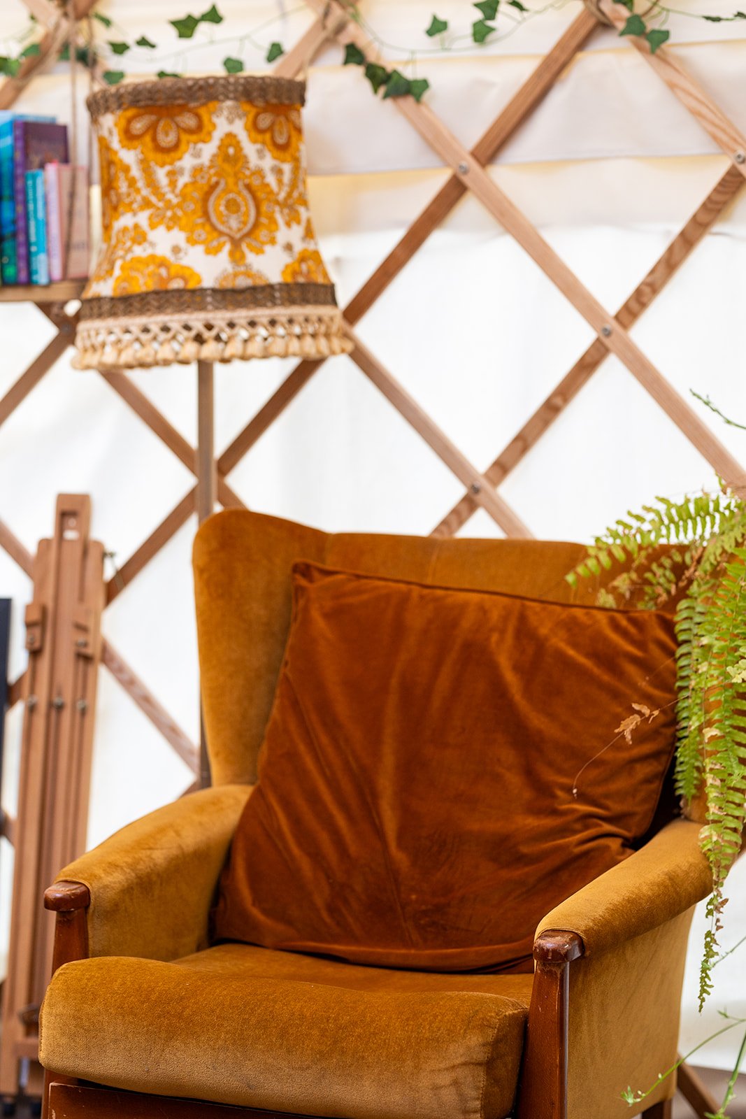 A cozy reading nook featuring a vintage mustard yellow armchair with a large rust-colored velvet pillow, a tall floor lamp with a detailed yellow, white, and brown lampshade, a wooden lattice wall, some green foliage, and a small bookshelf with color