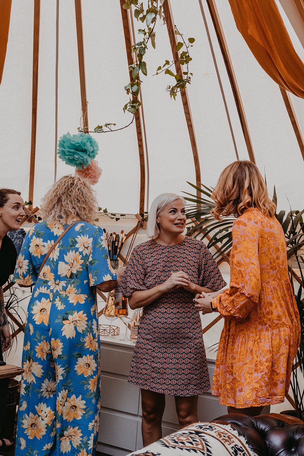 Four women talking at an indoor gathering, some wearing colorful, patterned dresses, with a tentlike ceiling and green plants in the background.