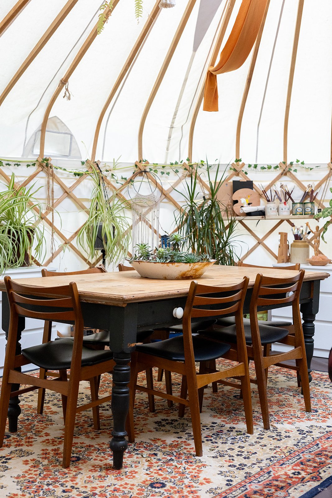 A cozy indoor space with a wooden dining table surrounded by five chairs, a decorative bowl with succulents on the table, a patterned rug underneath, and a lattice-style wall with hanging plants and art supplies in the background.