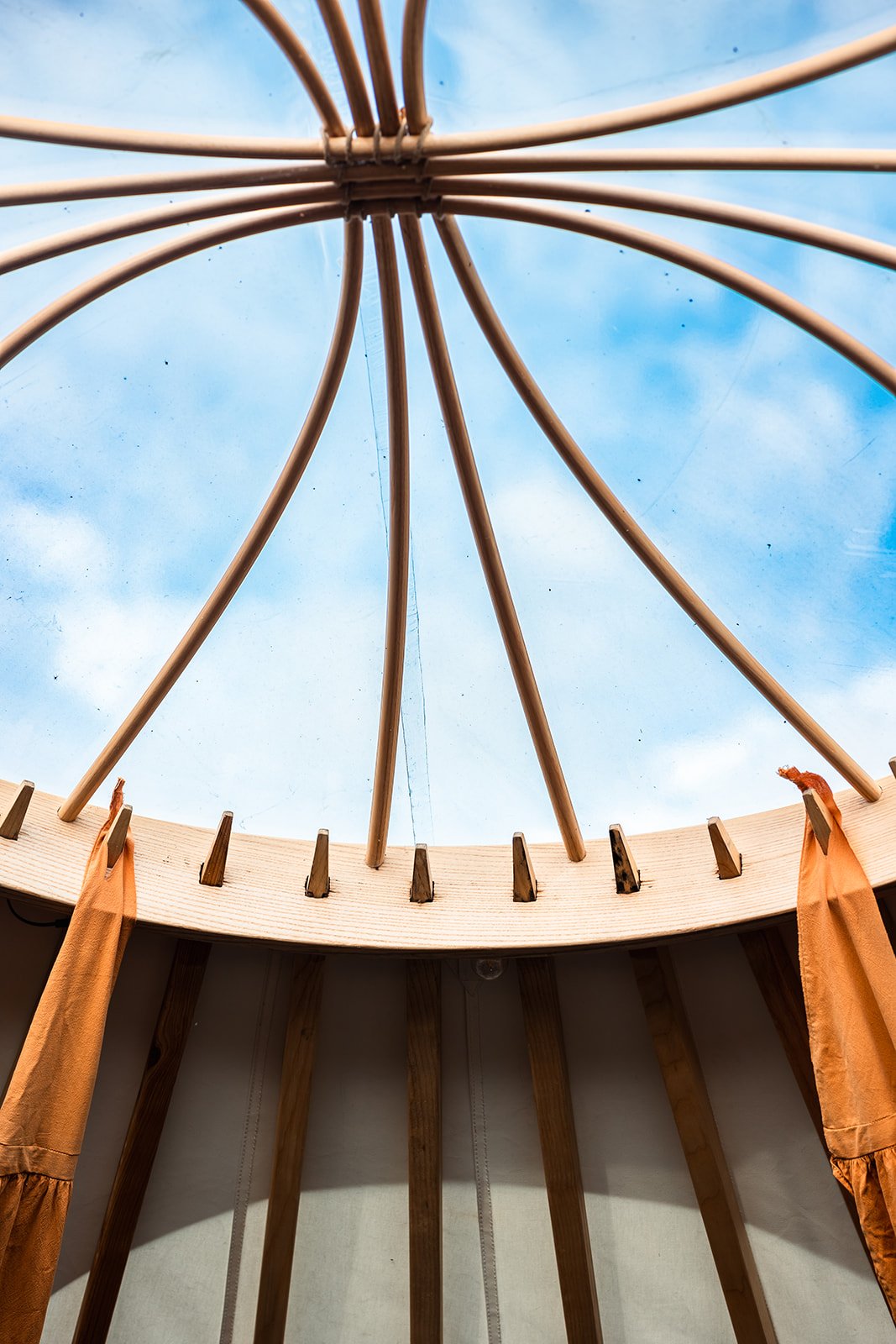 Interior view of a round structure with wooden beams and orange curtains, looking up at a blue sky through the open roof.