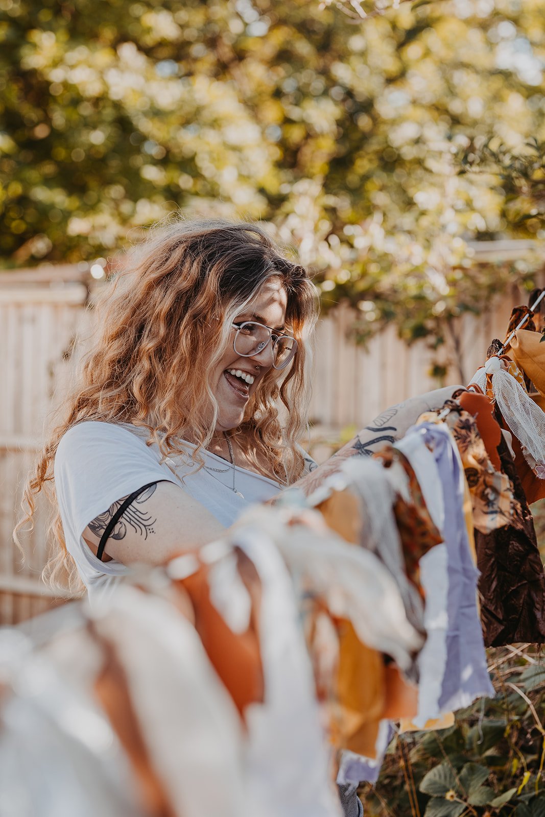 Woman with curly hair, glasses, and tattoos smiling outdoors near a fence, with various pieces of fabric or cloth hanging in front of her, possibly in a park or backyard.