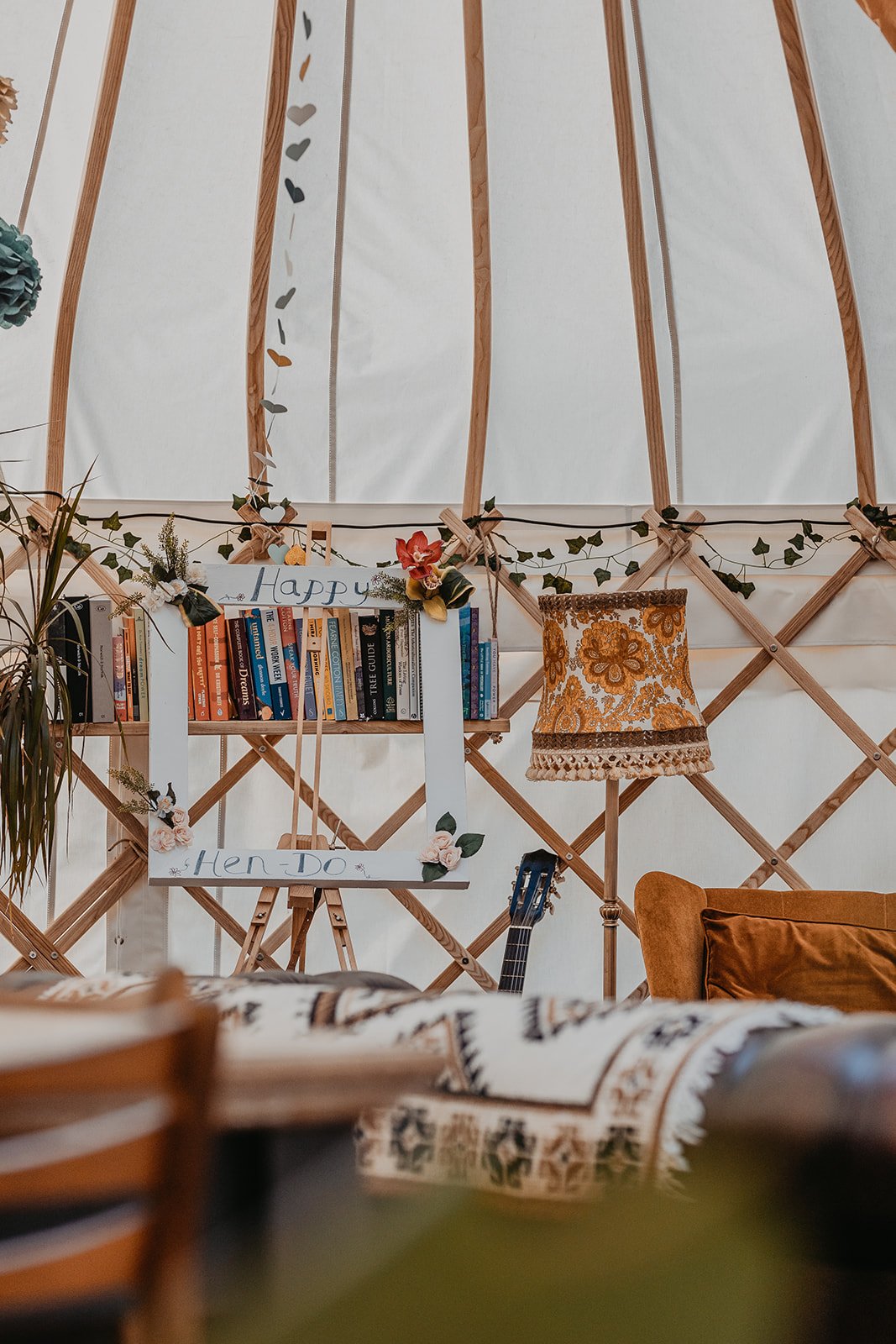 Interior of a yurt with a bookshelf, decorative lighting, a sign that says 'Happy 1st Hen Do', a guitar, a window with floral-patterned fabric, and a cozy armchair.