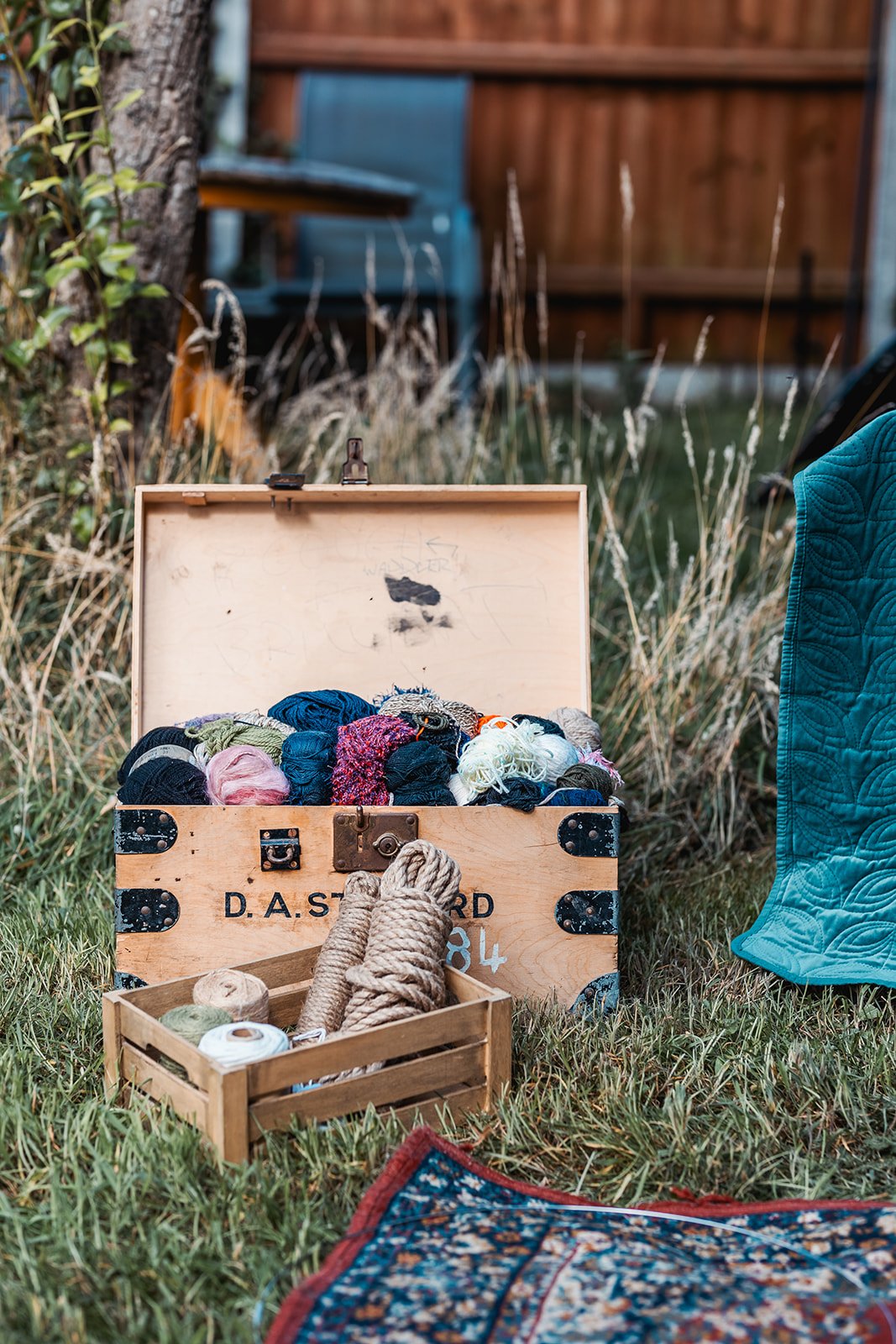 Open wooden box filled with colorful yarns and threads, and a small wooden crate with spools of twine, set outdoors on grass near a blue quilt and a tree.