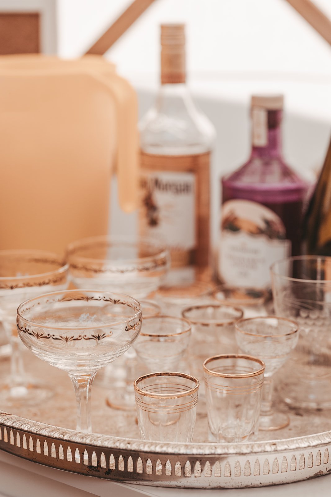 A collection of vintage glassware with intricate gold designs on a silver tray, along with bottles of liquor in the background.
