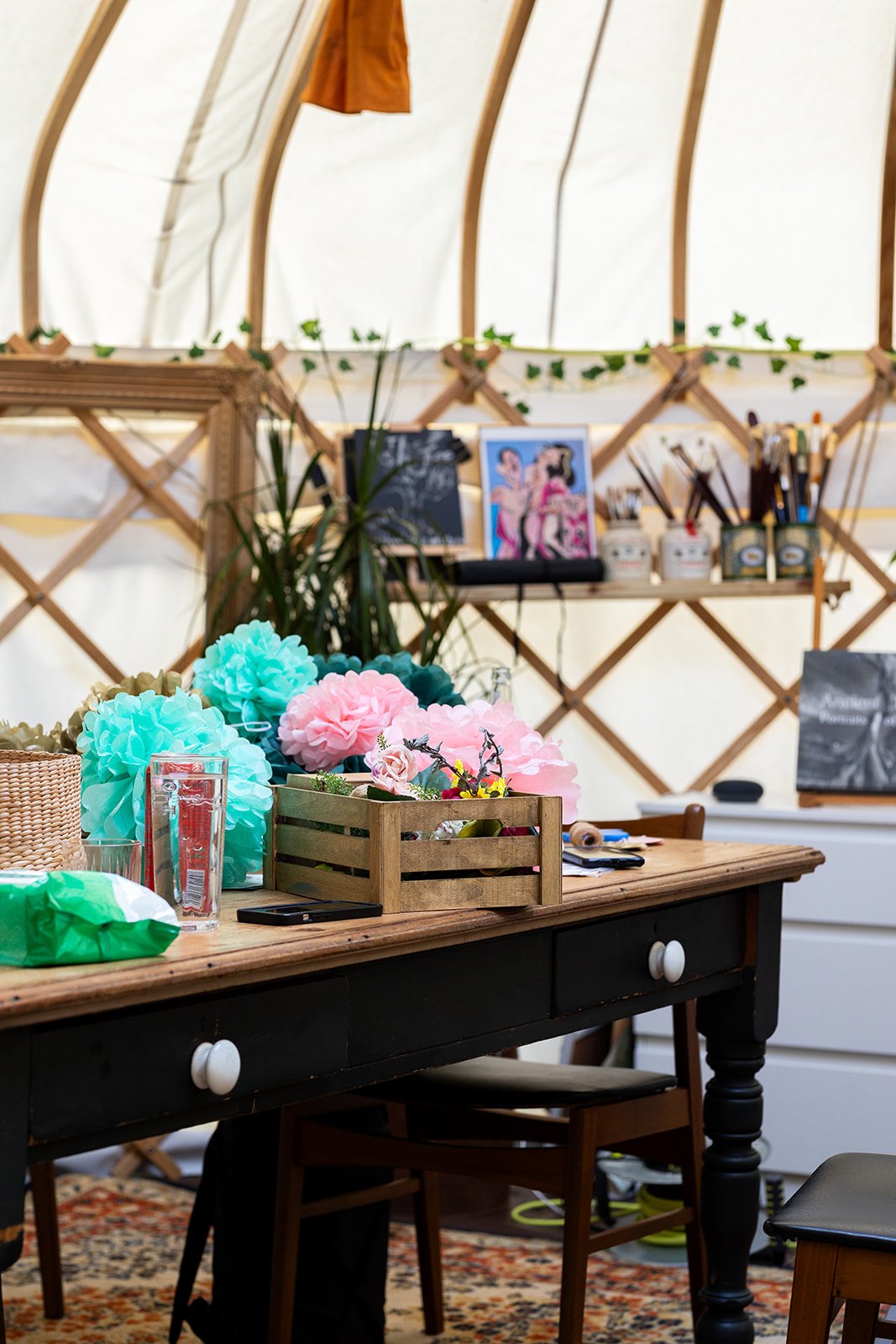 A crafting or decoration workspace with a table holding colorful paper pom-poms, flowers, a glass of water, a phone, and craft supplies. Behind the table is a wooden shelf with brushes, jars, and framed artwork. The background features a white canvas