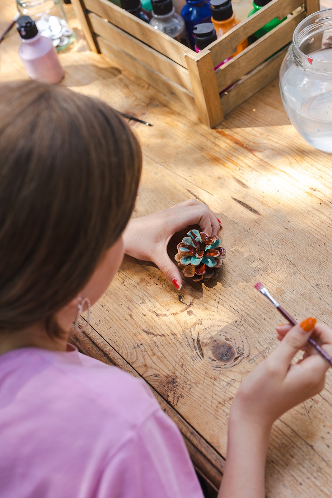 A person painting a pinecone on a wooden table with a small brush, with a bottle of paint and a glass water bowl nearby.