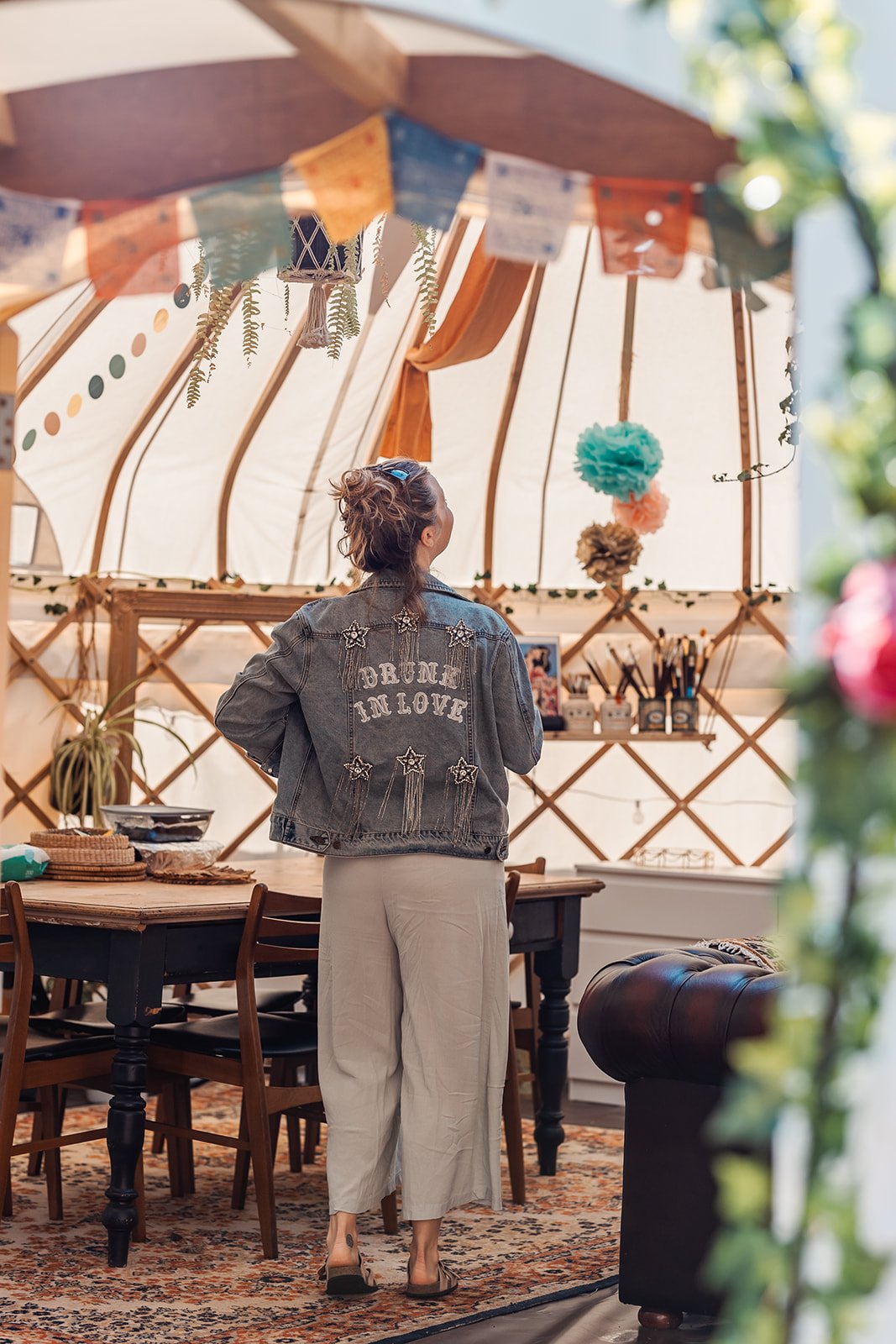 A woman stands inside a decorated tent, looking away. She wears a denim jacket with the words "DRUNK IN LOVE" on the back, beige wide-leg pants, and sandals. The tent is adorned with colorful garlands, hanging paper pom-poms, and plants, set up over